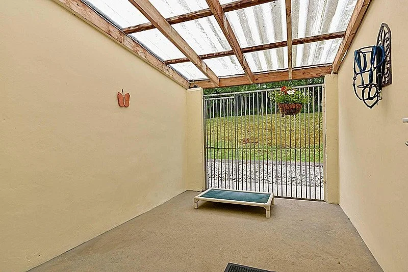 Enclosed patio with beige walls, a flat pet bed, a hanging flower basket, a decorative wall piece, and a metal gate leading to a grassy yard with trees.