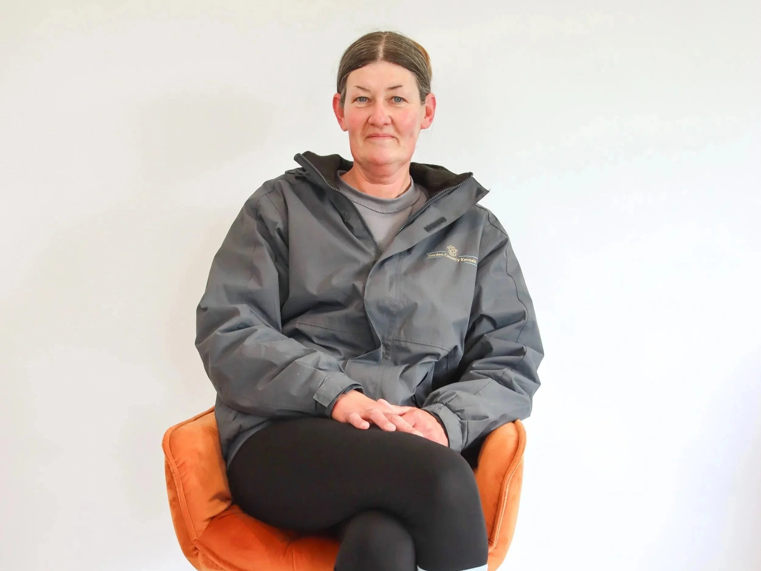 A middle-aged woman with short brown hair sitting in an orange chair against a plain white wall, wearing a gray jacket and black pants.