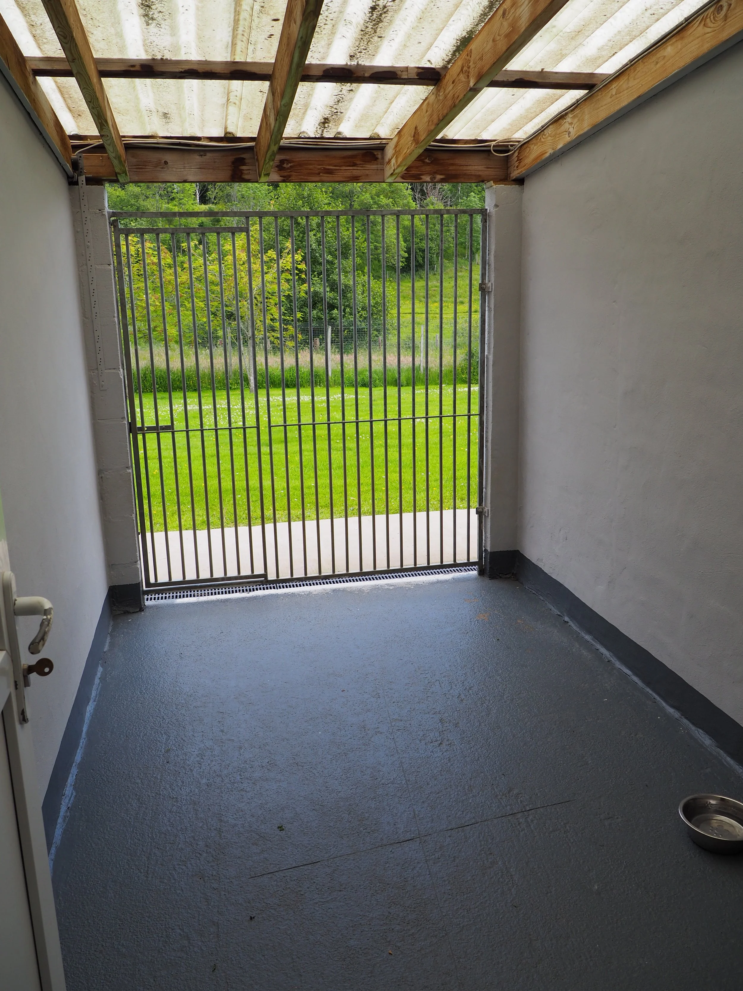 A small enclosed porch or patio area with a concrete floor, bordered by white walls. At the back of the area, there is a metal security gate leading to a grassy yard with trees and a fence in the distance. The ceiling of the porch has exposed wooden 