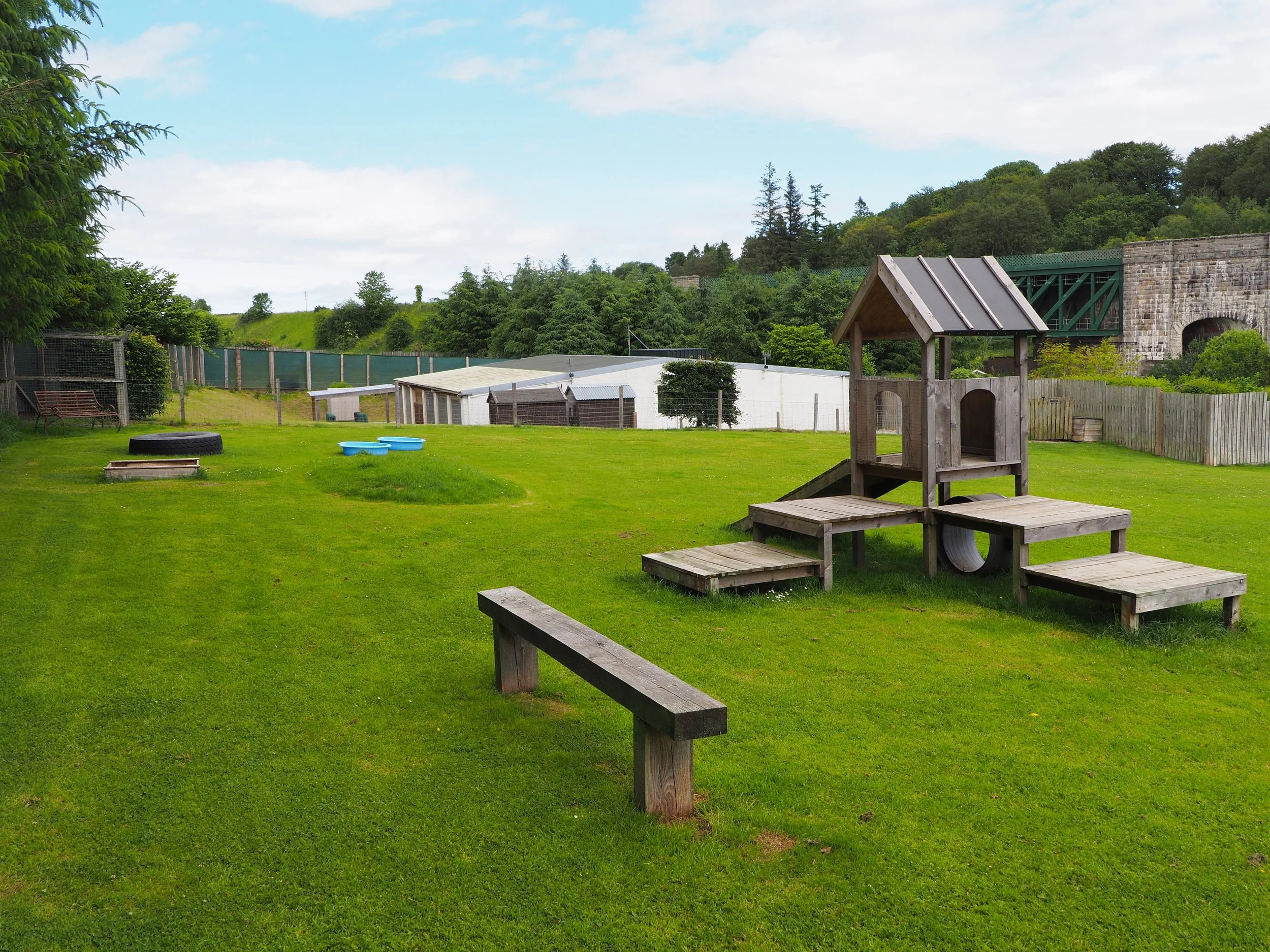 A grassy area with a play structure that has a slide, surrounded by fencing with trees and shrubs, a building with a slanted roof, and an old stone bridge in the background.