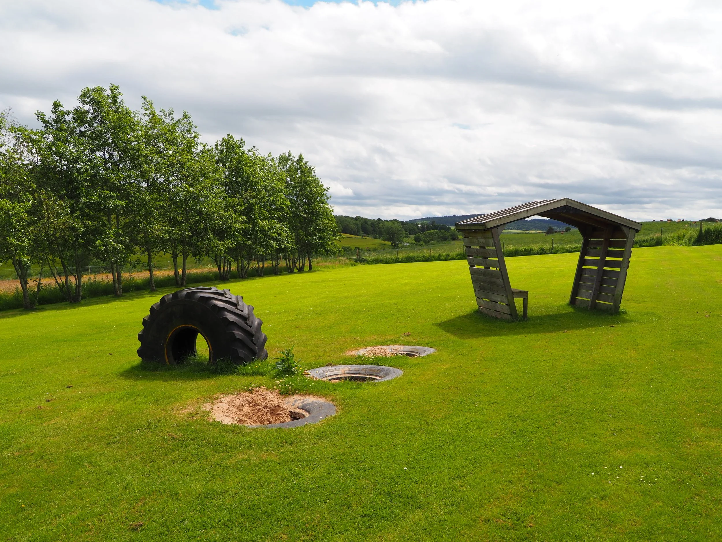 Open manhole covers on a grassy field with a wooden bench and trees in the background under a cloudy sky.