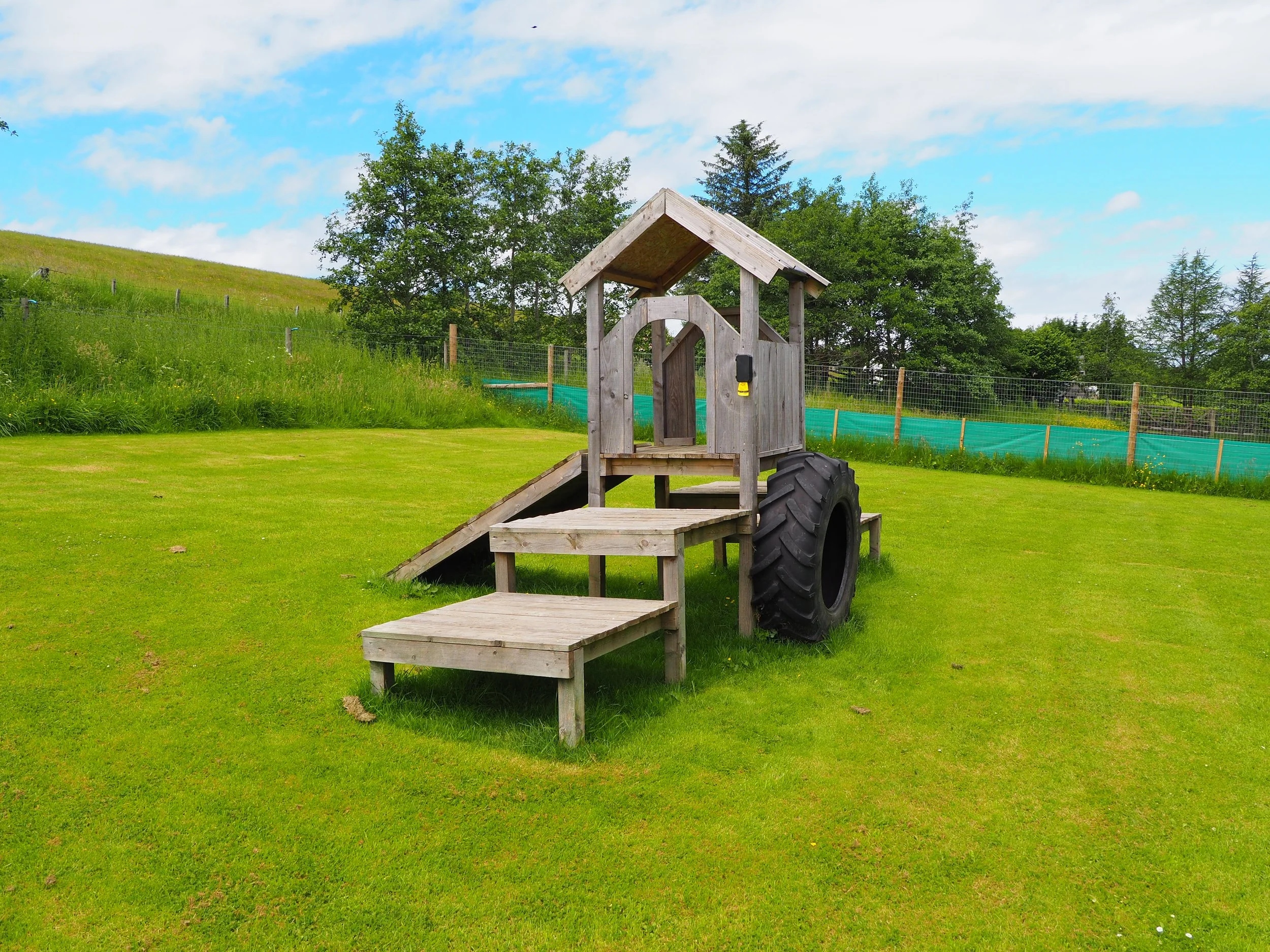 A wooden play structure with a ramp, small roof, and steering wheel in an outdoor grassy area, with green trees, a fence, and a partly cloudy sky in the background.