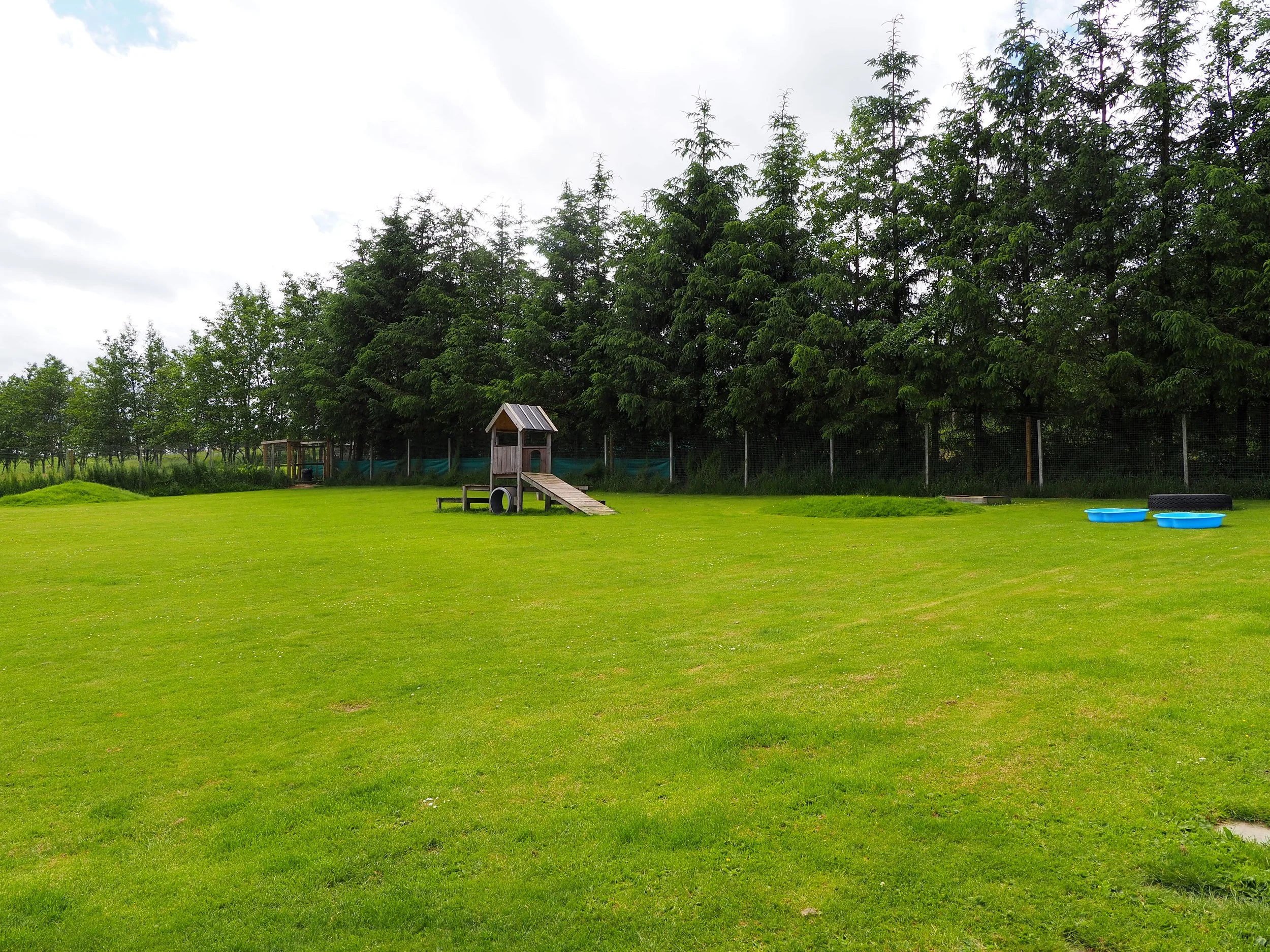 A playground with a small wooden playhouse and tunnel on a well-manicured grassy field, surrounded by trees and a fence, overcast sky.