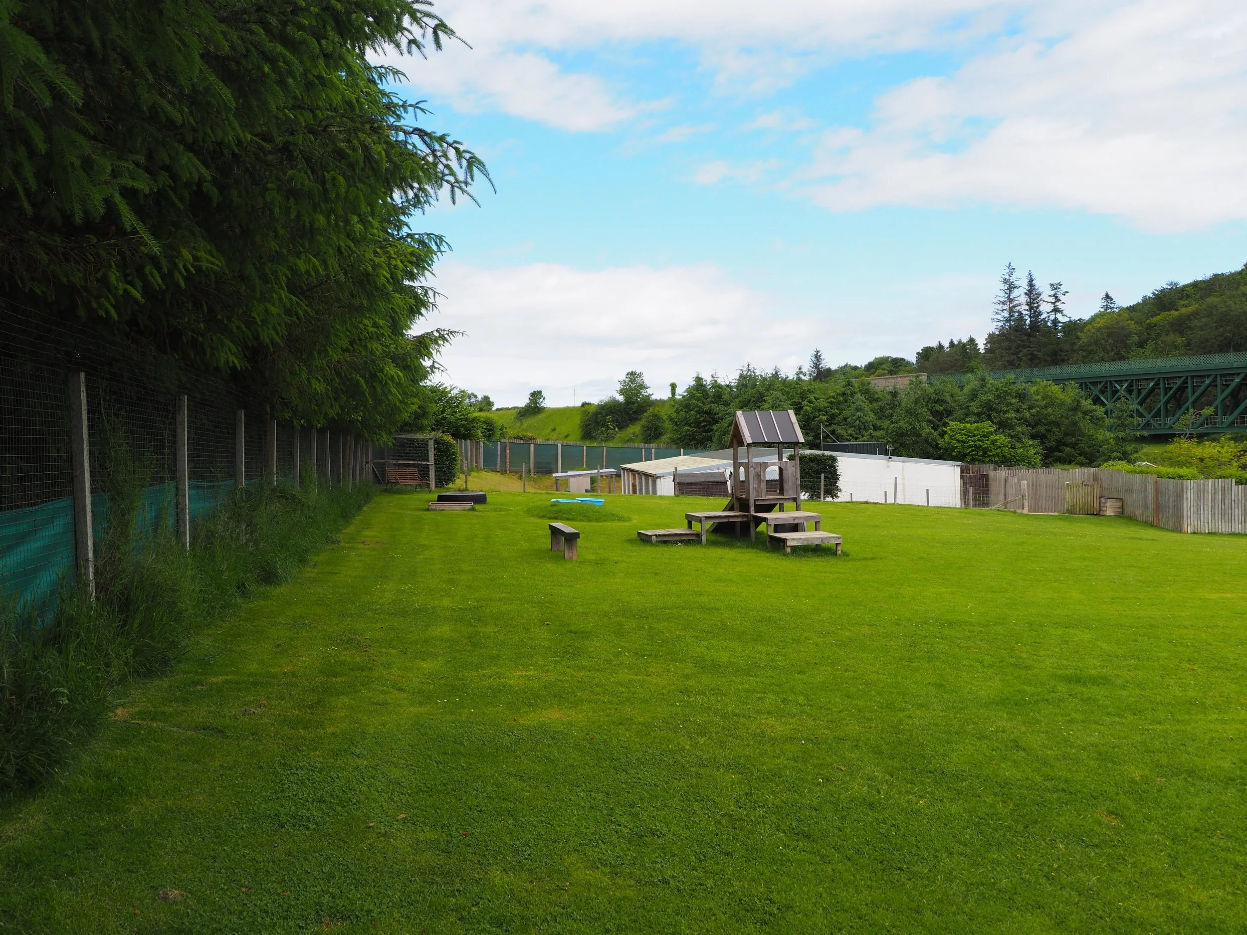 A green outdoor park with a grassy yard, benches, a small wooden playhouse, a tire, and a fence. In the background, there are trees, a hill, and an overpass bridge.