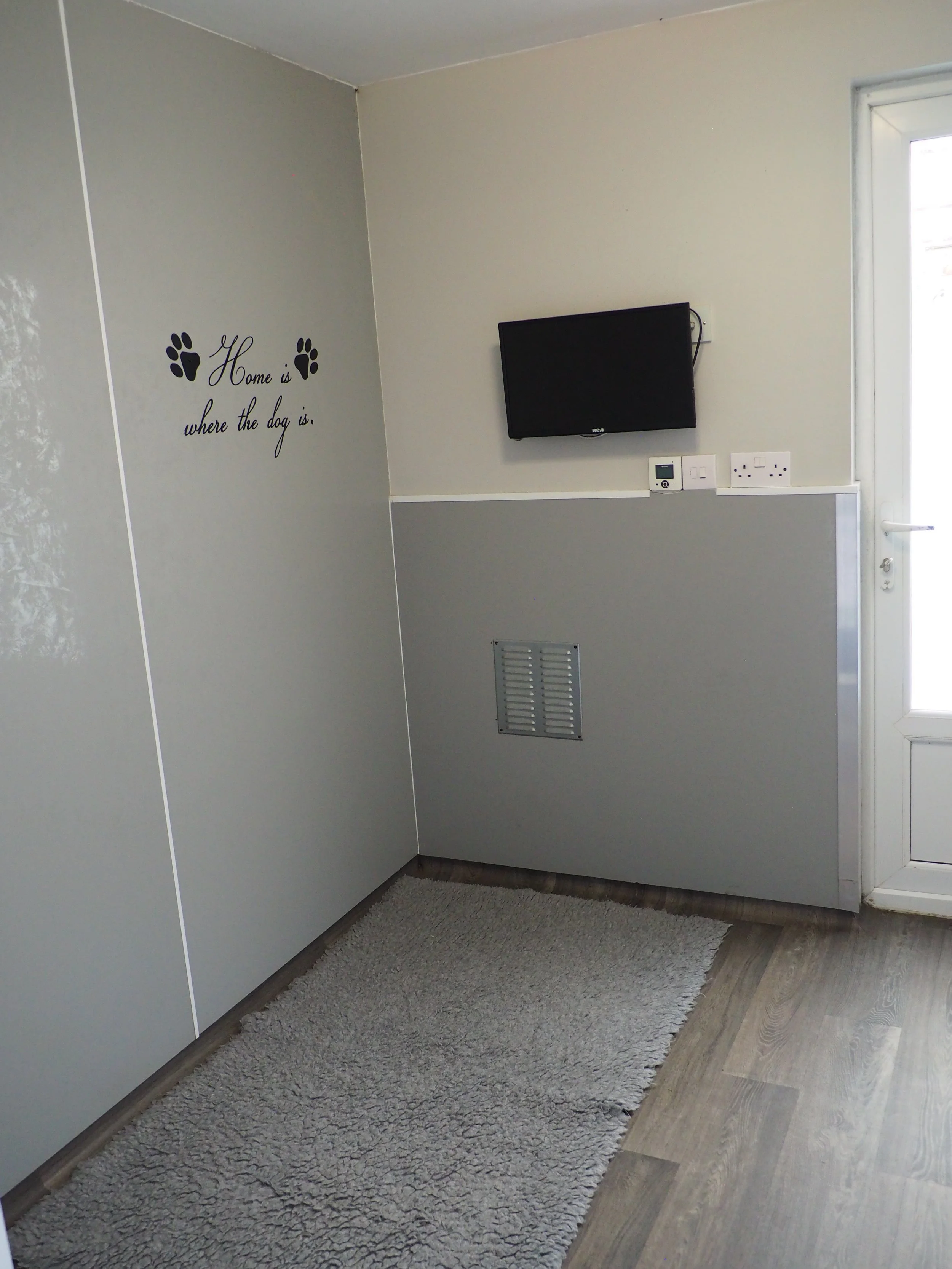 A corner of a room with light gray walls, featuring a quote sticker with paw prints that says 'Home is where the dog is.' There is a small wall-mounted monitor and electrical outlets, with a door on the right side letting in sunlight. A beige shaggy 