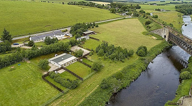 Aerial view of a rural area with houses, green fields, trees, a river, and a bridge.