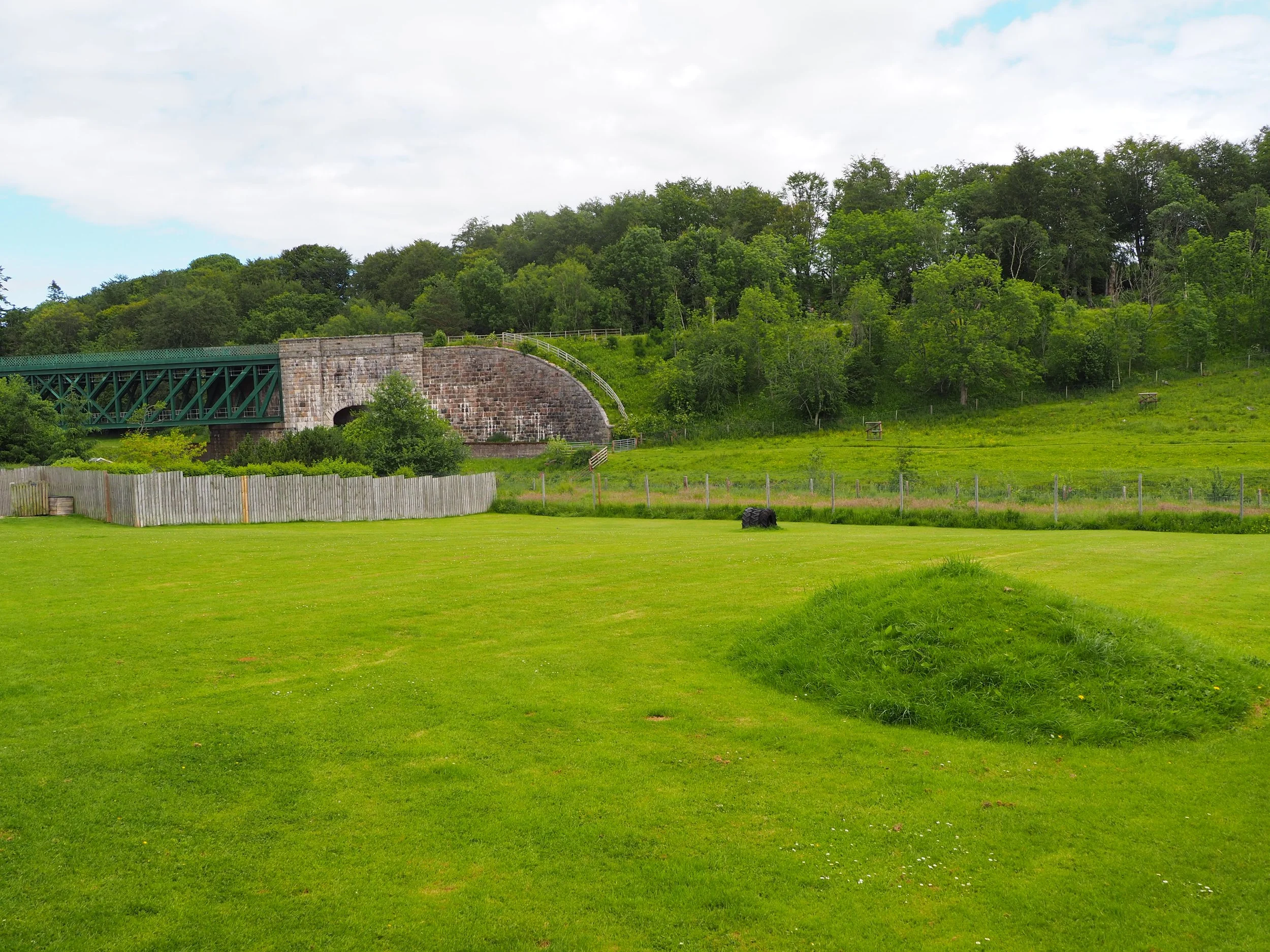 A lush green field with a wooded hill in the background and a stone bridge crossing from left to right in the middle ground. There is a white picket fence and some small bushes in the foreground, with a partly cloudy sky overhead.