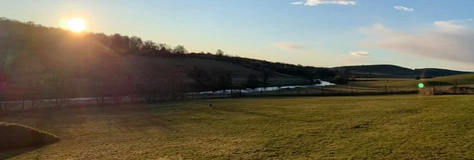 Sunset over a rural landscape with rolling hills, a river, and a grassy field.
