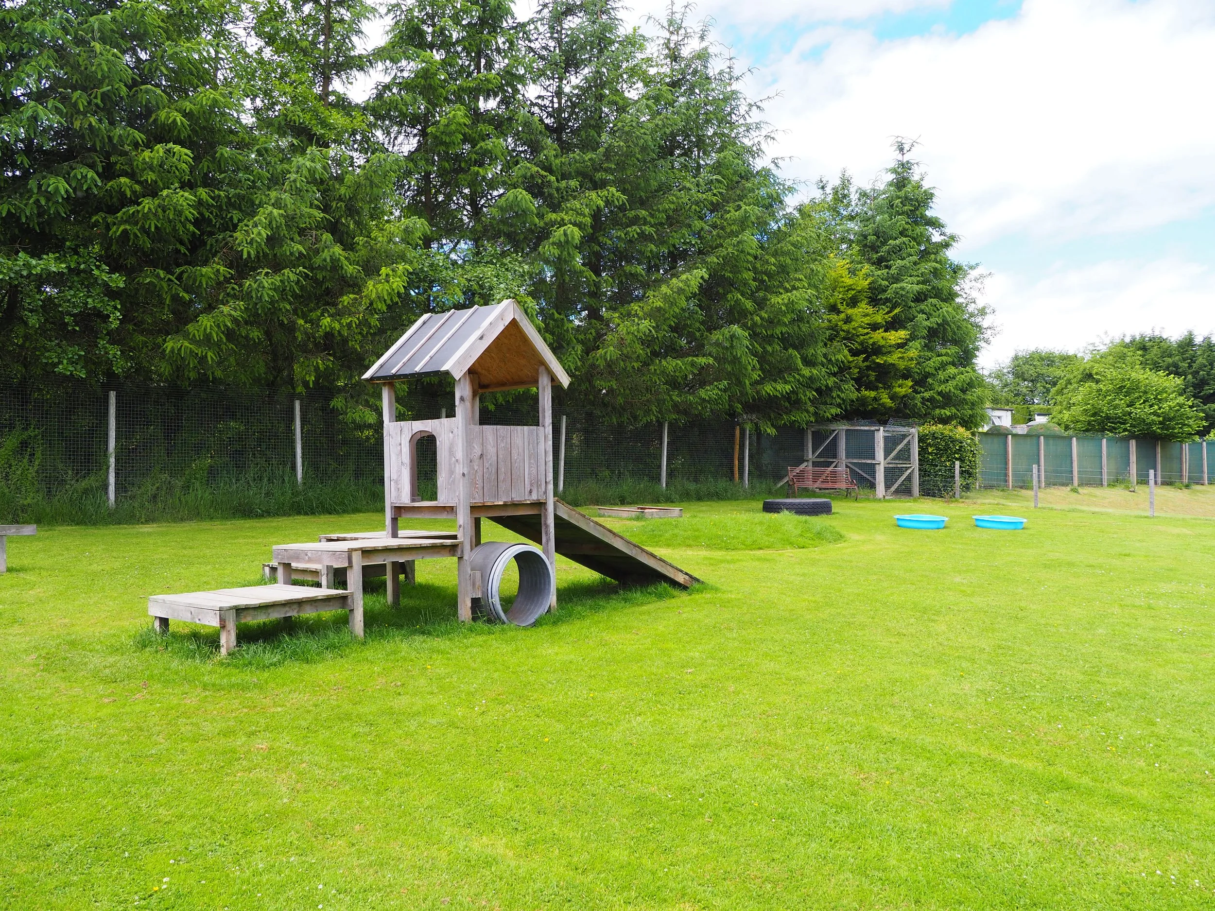 A grassy backyard with a small wooden play structure, two blue plastic pools, a children's slide, and a tire, with tall green trees and a fence in the background.