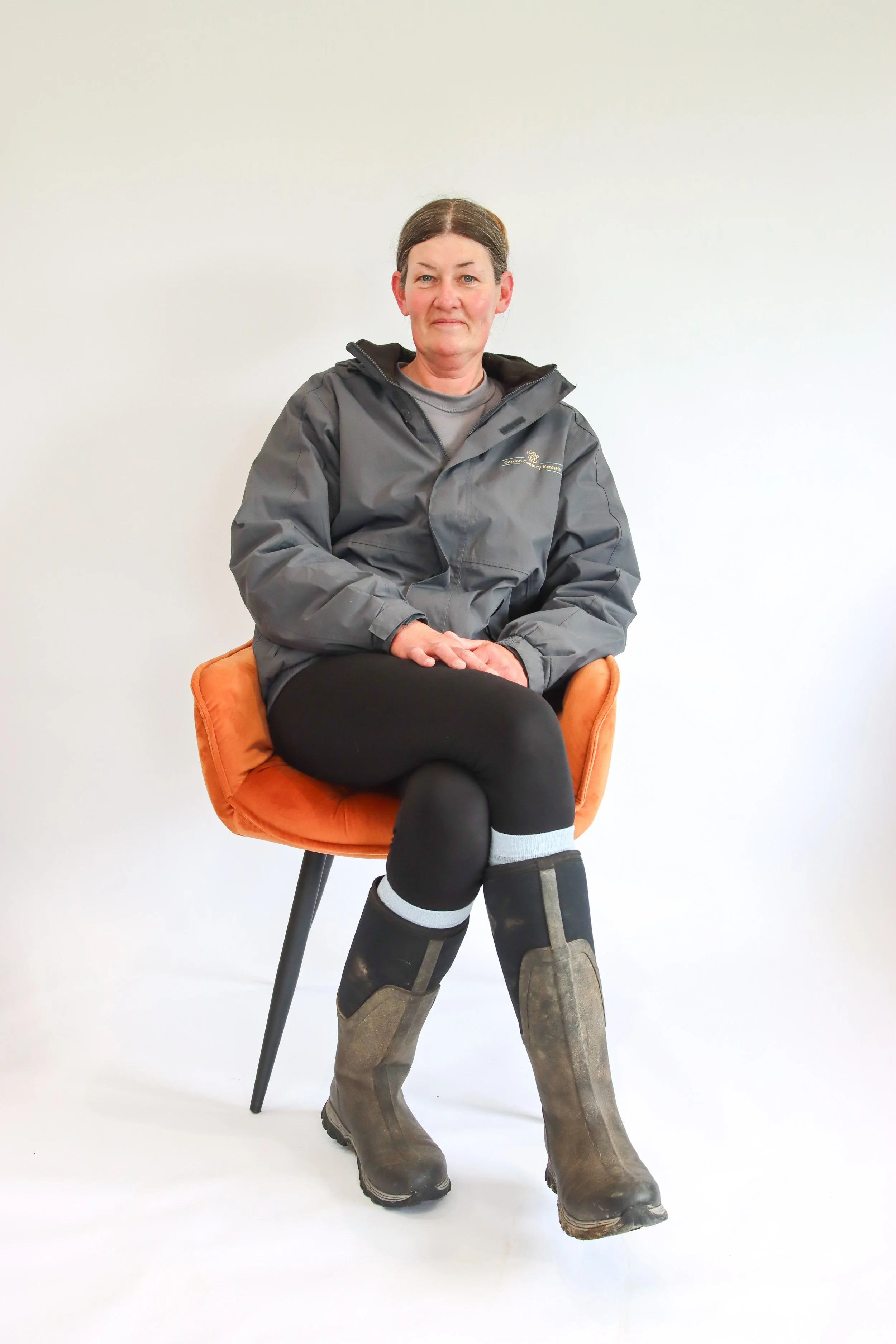 Woman sitting on an orange chair wearing a gray jacket, black leggings, and brown boots, against a white background.