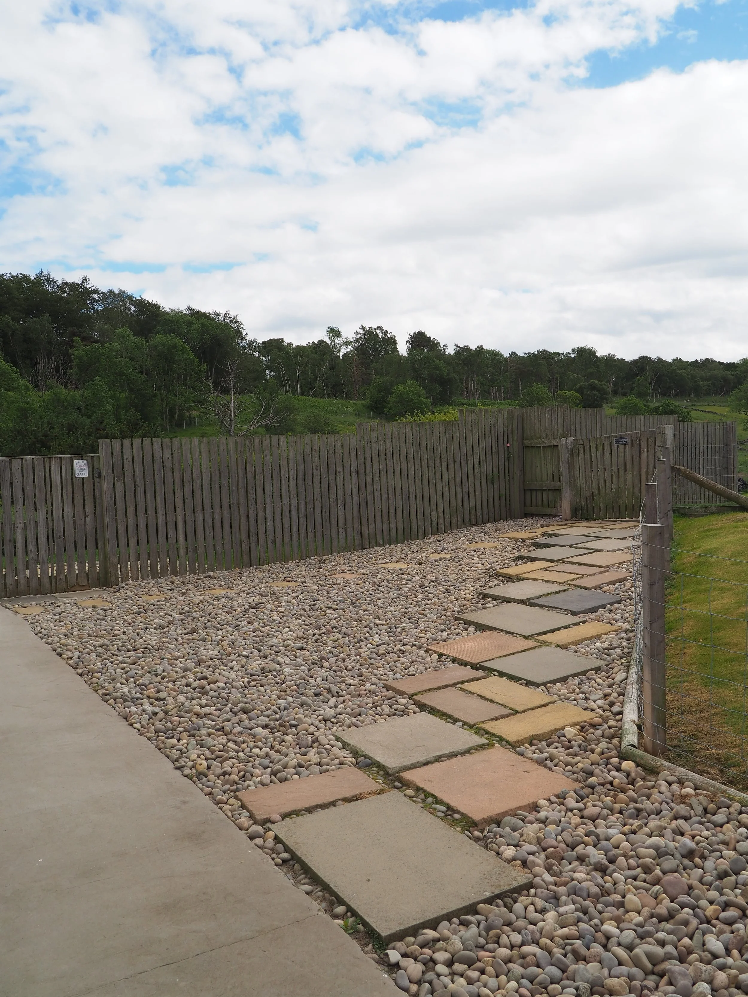 A pathway with large square concrete pavers next to a bed of small rounded rocks, bordered by a wooden fence with trees and hills in the background under a partly cloudy sky.