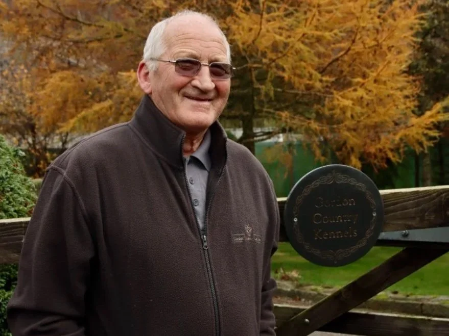 An older man with glasses and a gray hat smiling outdoors in front of a wooden fence and autumn trees. There is a sign that reads "Cashion County K-9s."