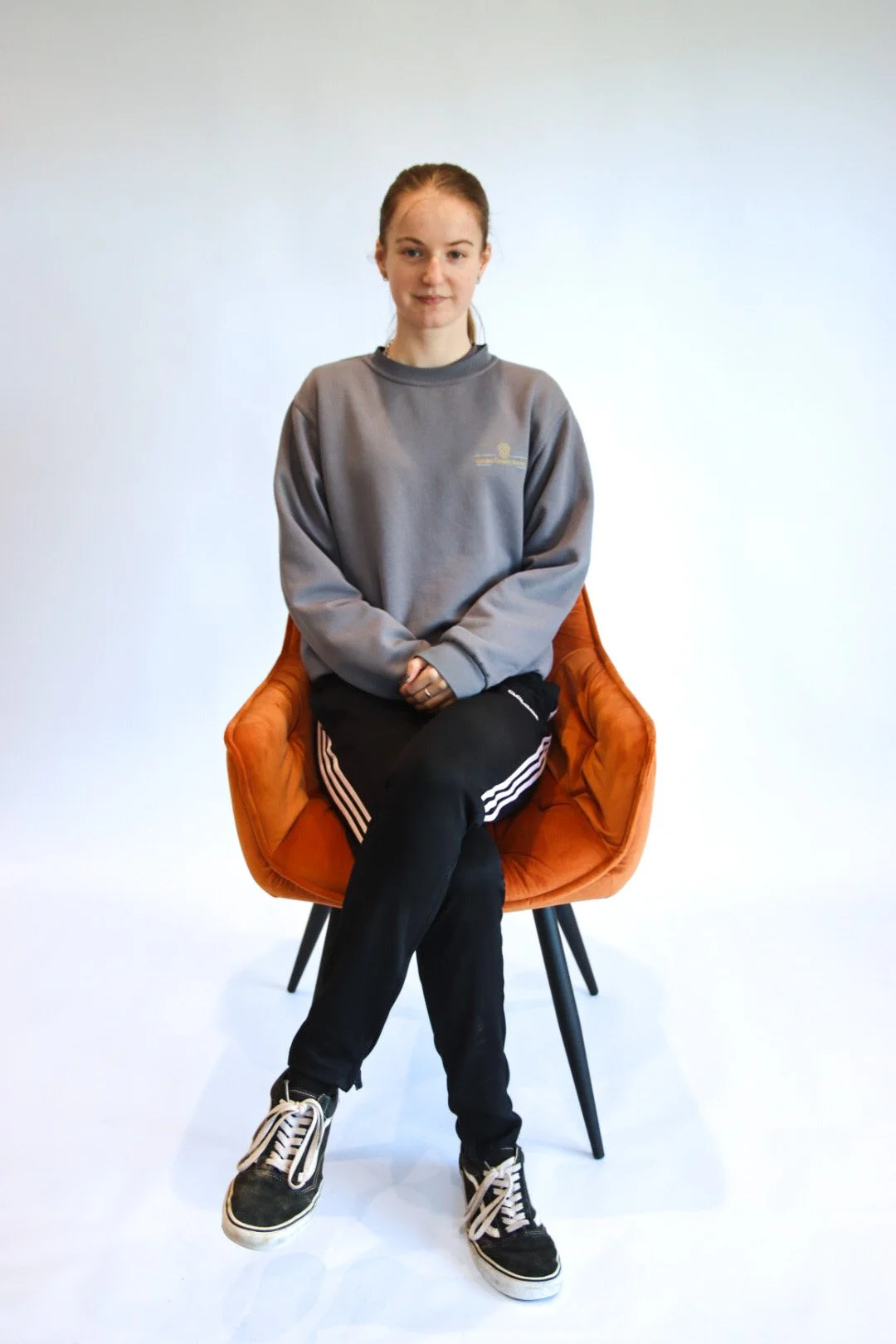 A young woman with red hair sitting cross-legged on an orange chair with black legs, wearing a gray sweatshirt, black Adidas track pants, and black Vans sneakers.
