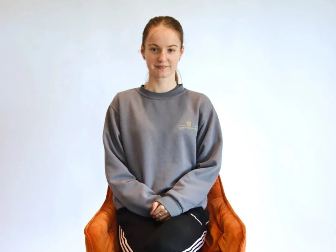 A young woman with red hair sitting on an orange chair against a plain white background, wearing a gray sweatshirt and black pants.