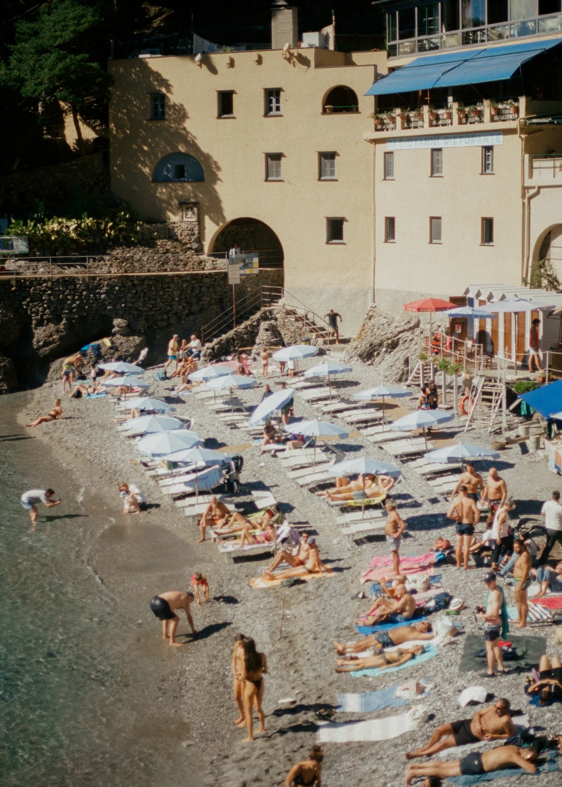 People relaxing and sunbathing on a pebbled beach with umbrellas, near a building with outdoor seating and a staircase leading to the shore.