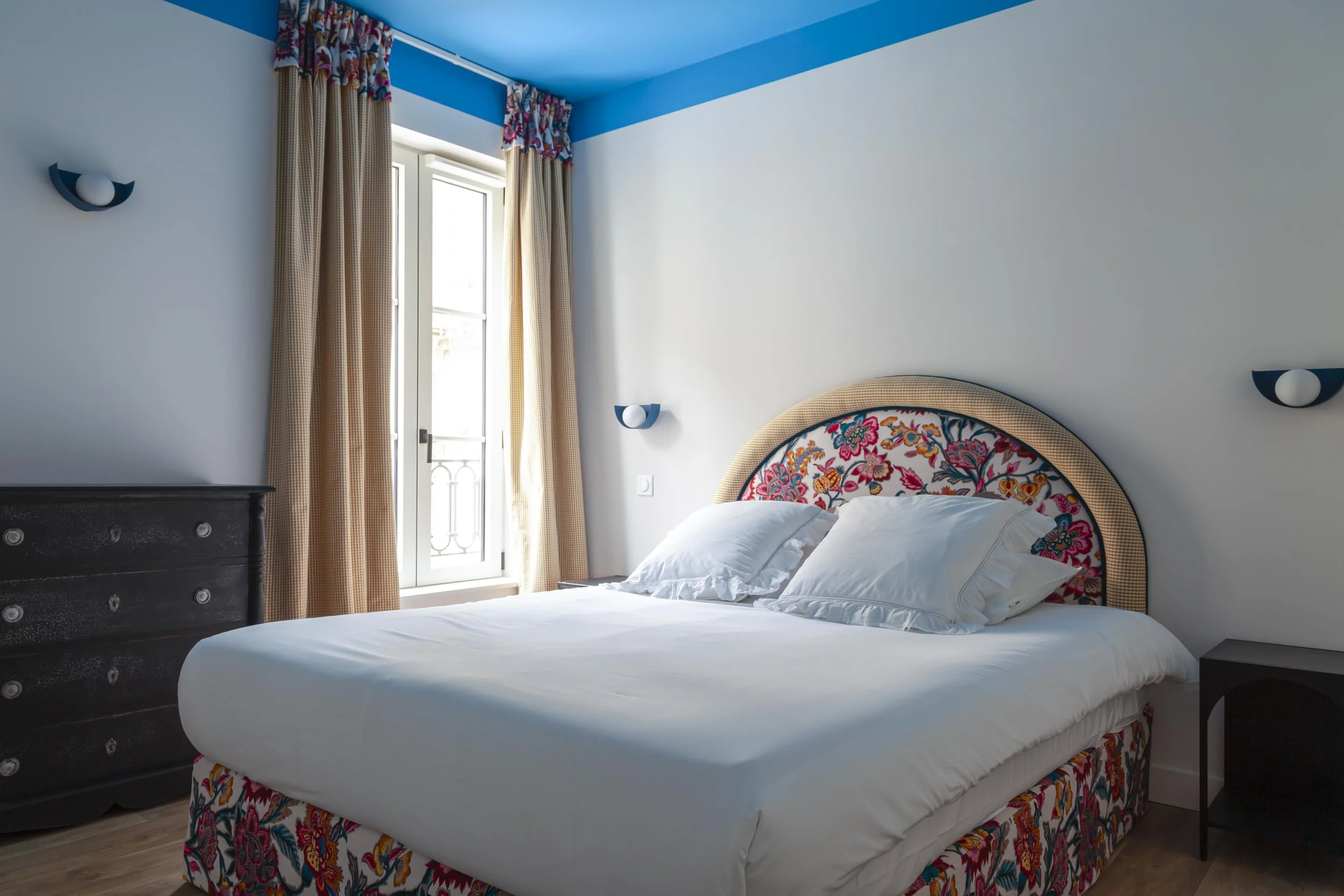 Chambre avec lit double, têtes de lit colorées avec motifs floraux, rideaux beige avec détails colorés, deux lampes murales, fenêtre avec porte-fenêtre, armoire noire en bois, mur blanc, plafond bleu, sol en bois.