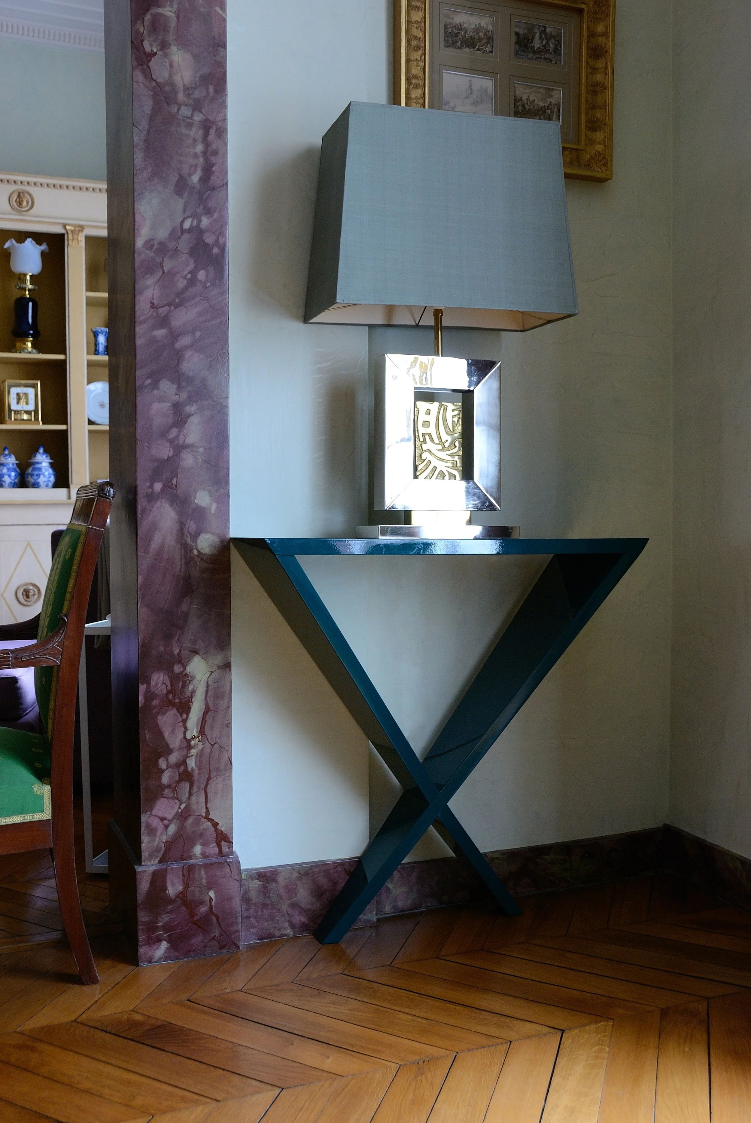 Intérieur d'une pièce avec une lampe de table, un tableau dans un cadre doré et un meuble en forme de V avec un miroir, sur un sol en parquet.