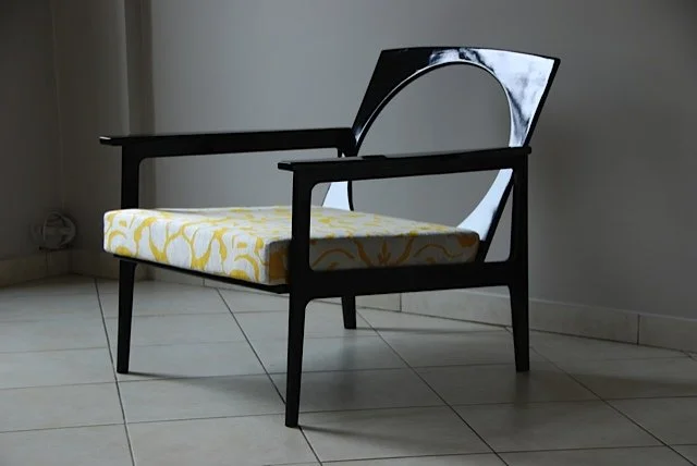 Fauteuil en bois noir avec coussin à motifs jaunes et blancs dans une pièce avec sol en carreaux et mur gris.