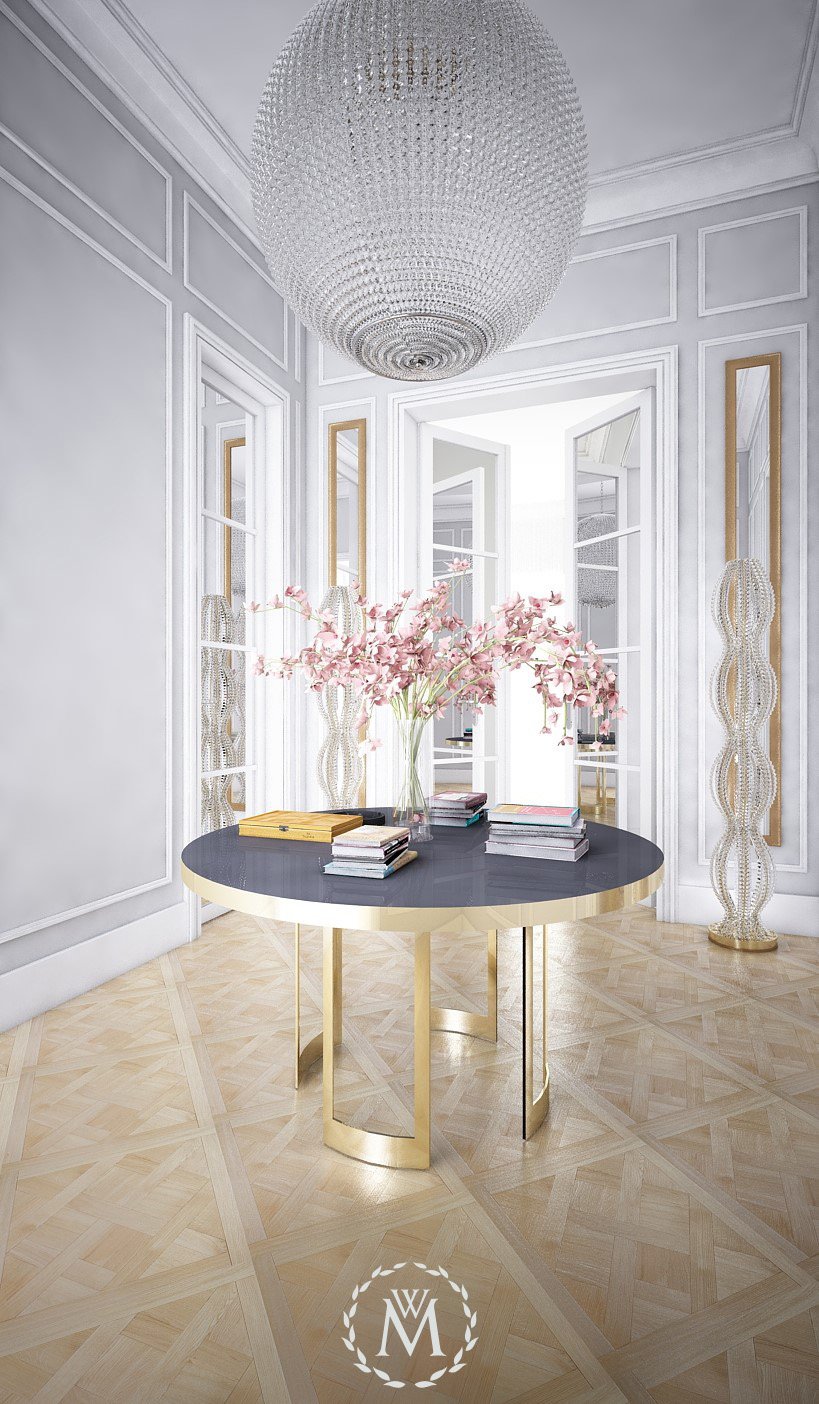Intérieur d'une pièce élégante avec une table ronde noire et or, décorée de livres et de fleurs roses dans un vase. Sur le plafond, un grand lustre en cristal, et dans le coin, une sculpture en forme de colonne. La pièce est lumineuse avec des murs blancs et des fenêtres grandes ouvertes.
