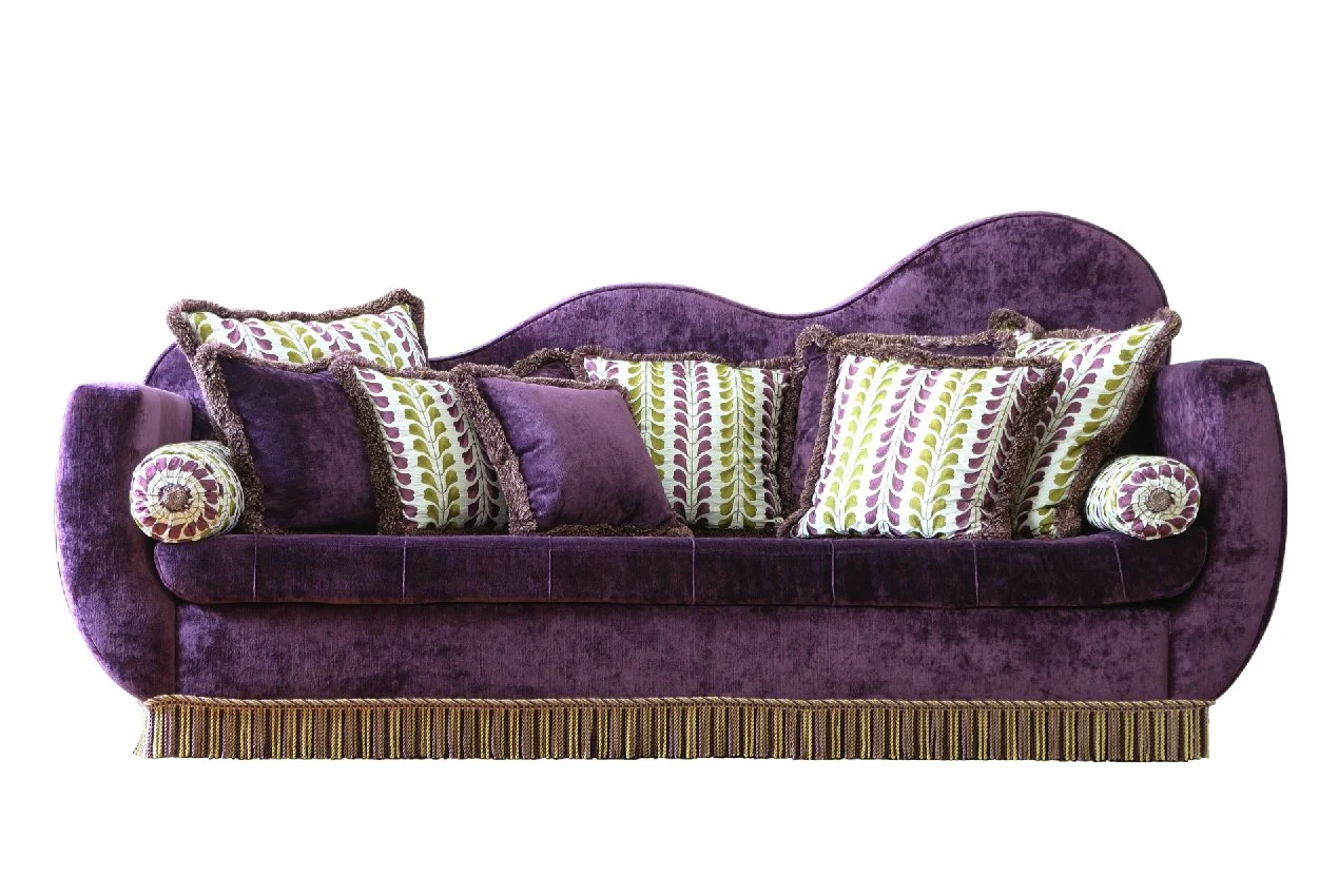 Un canapé violet en velours avec plusieurs coussins colorés et décoratifs, certains à motifs de feuilles vertes et lavandes, d'autres en velours violet, sur un fond blanc.