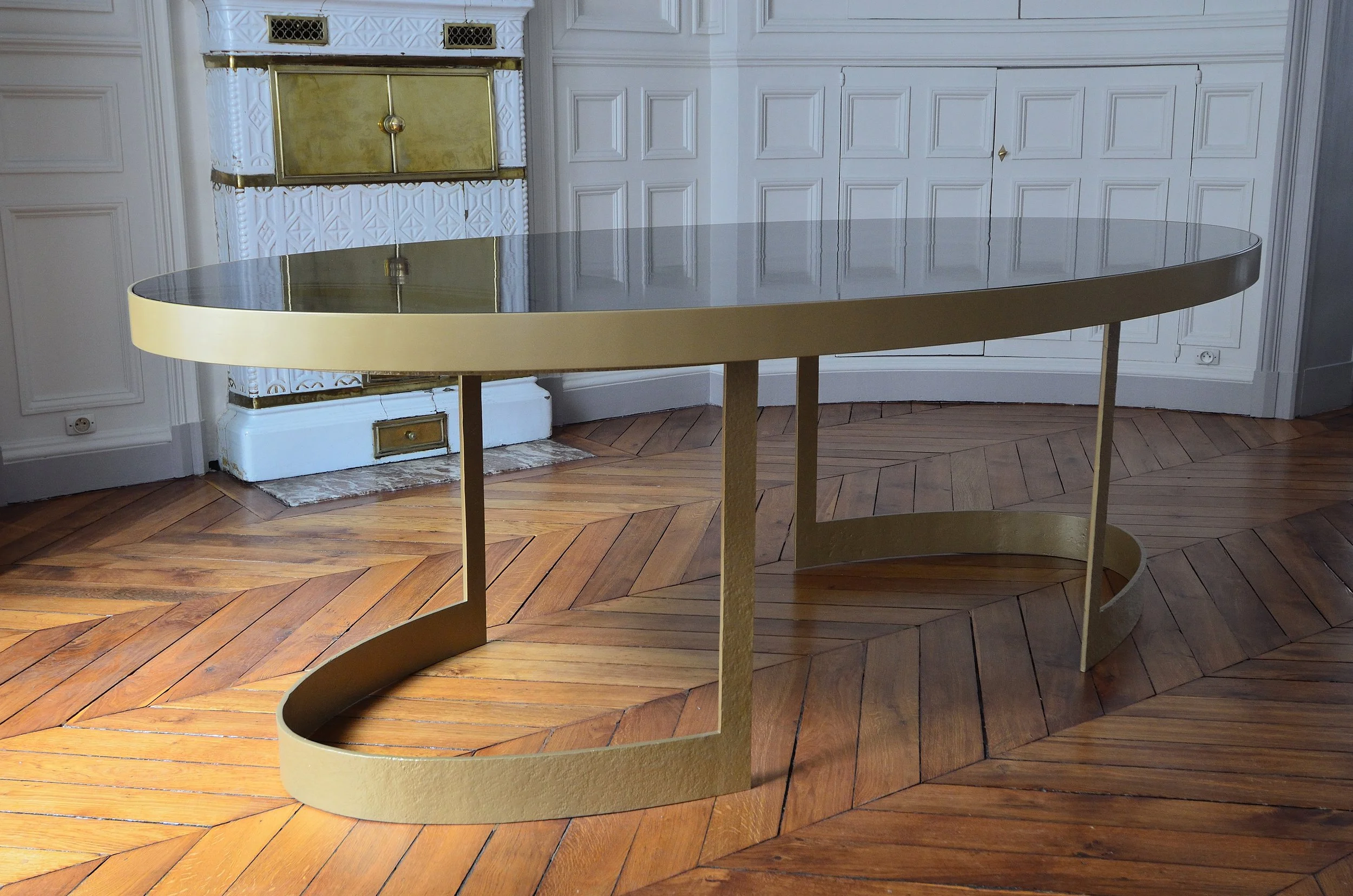 Une table moderne avec un dessus en verre foncé et une base en métal doré, placée dans une pièce avec un parquet en bois et des murs ornés de moulures blanches, présence d'une cheminée avec des détails dorés en arrière-plan.