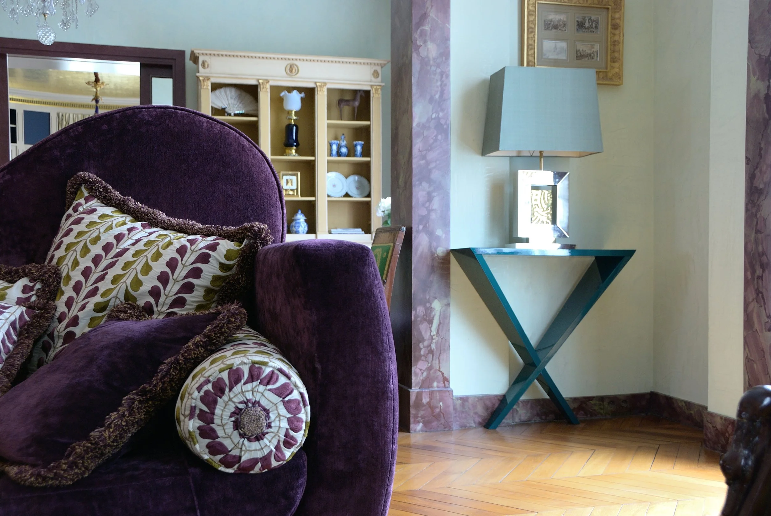 Intérieur d'un salon avec un canapé violet en velours, un coussin décoratif à motif floral, une étagère décorative avec des objets bleus et blancs, une lampe moderne avec abat-jour bleu, un tableau encadré au mur, et un sol en parquet en bois.