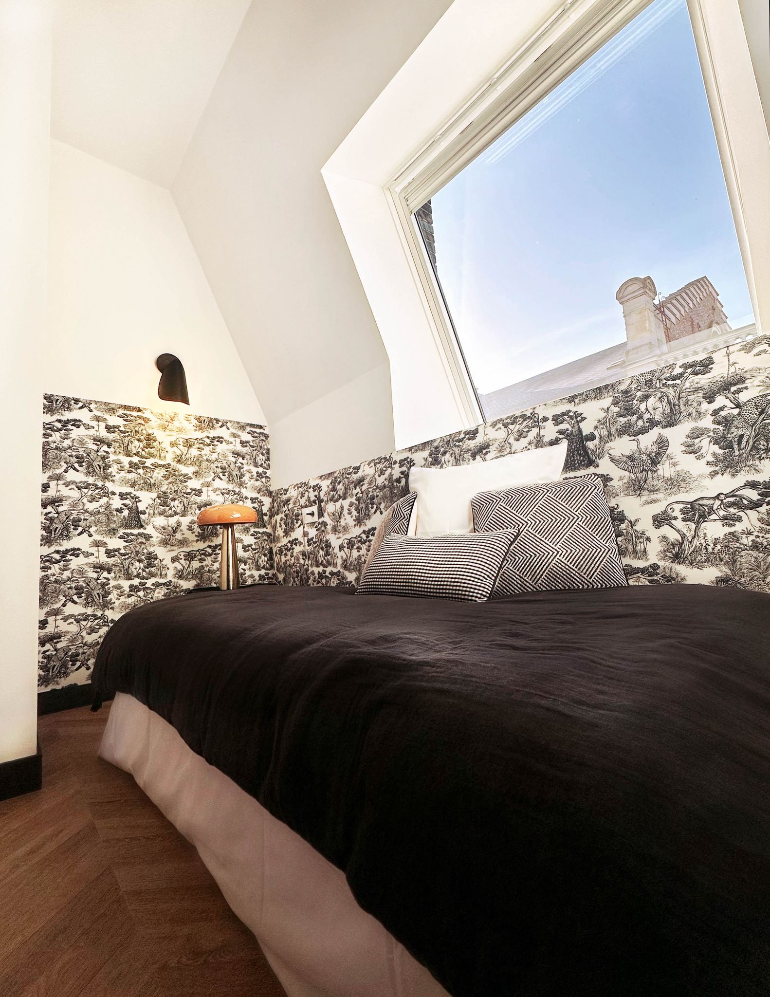 Une chambre avec un lit, des oreillers à motifs noirs et blancs, une tapisserie à motif animalier noir et blanc, une lampe dorée, un mur blanc avec une applique noire, une fenêtre donnant sur le ciel bleu avec des bâtiments en retrait, et un parquet 