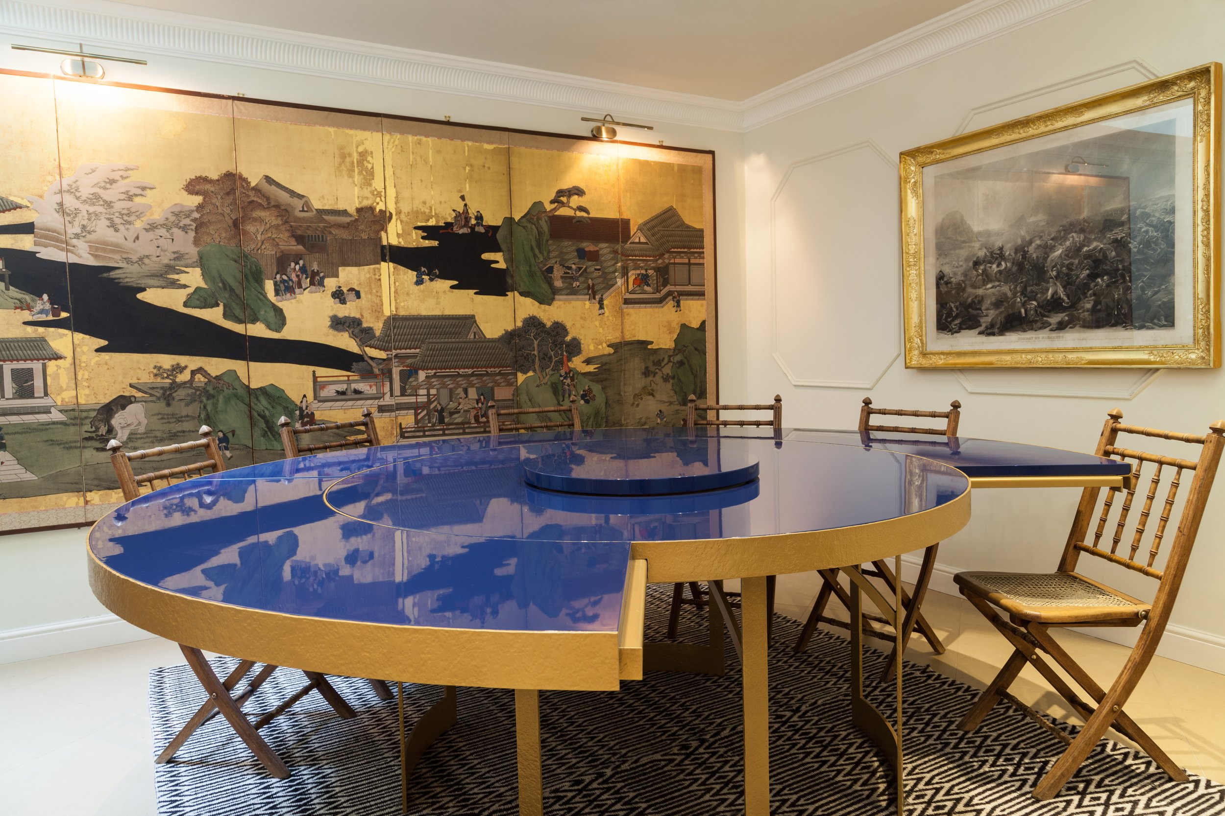 Une salle d'exposition avec une grande peinture japonaise ancienne accrochée au mur, un miroir encadré or et une table ronde bleue avec des chaises en bois autour, posée sur un tapis noir et blanc.