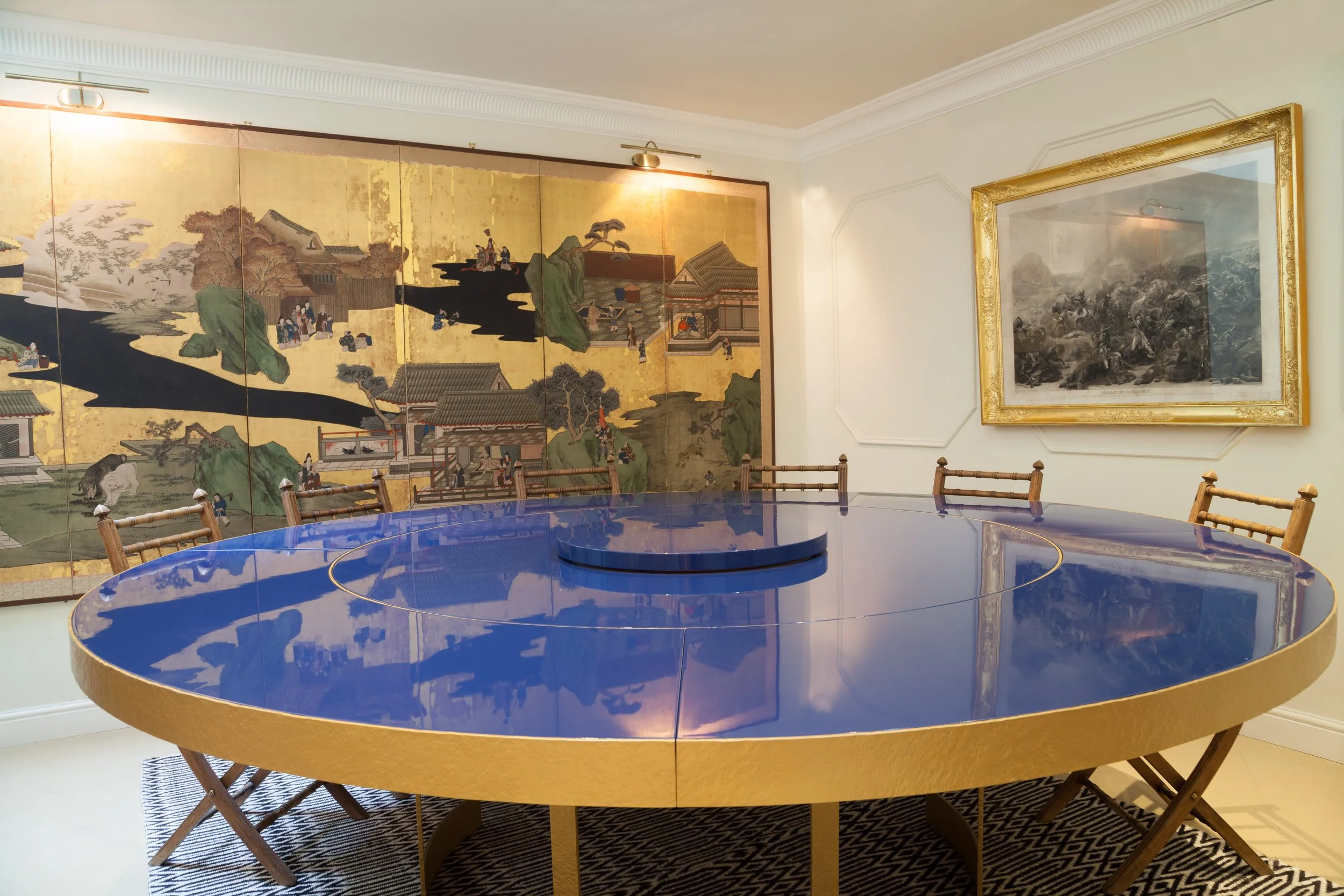Table ronde bleue brillante dans une salle d'exposition, avec des chaises en bois, derrière un mur décoré de peintures traditionnelles japonaises et de nombreux autres tableaux encadrés.