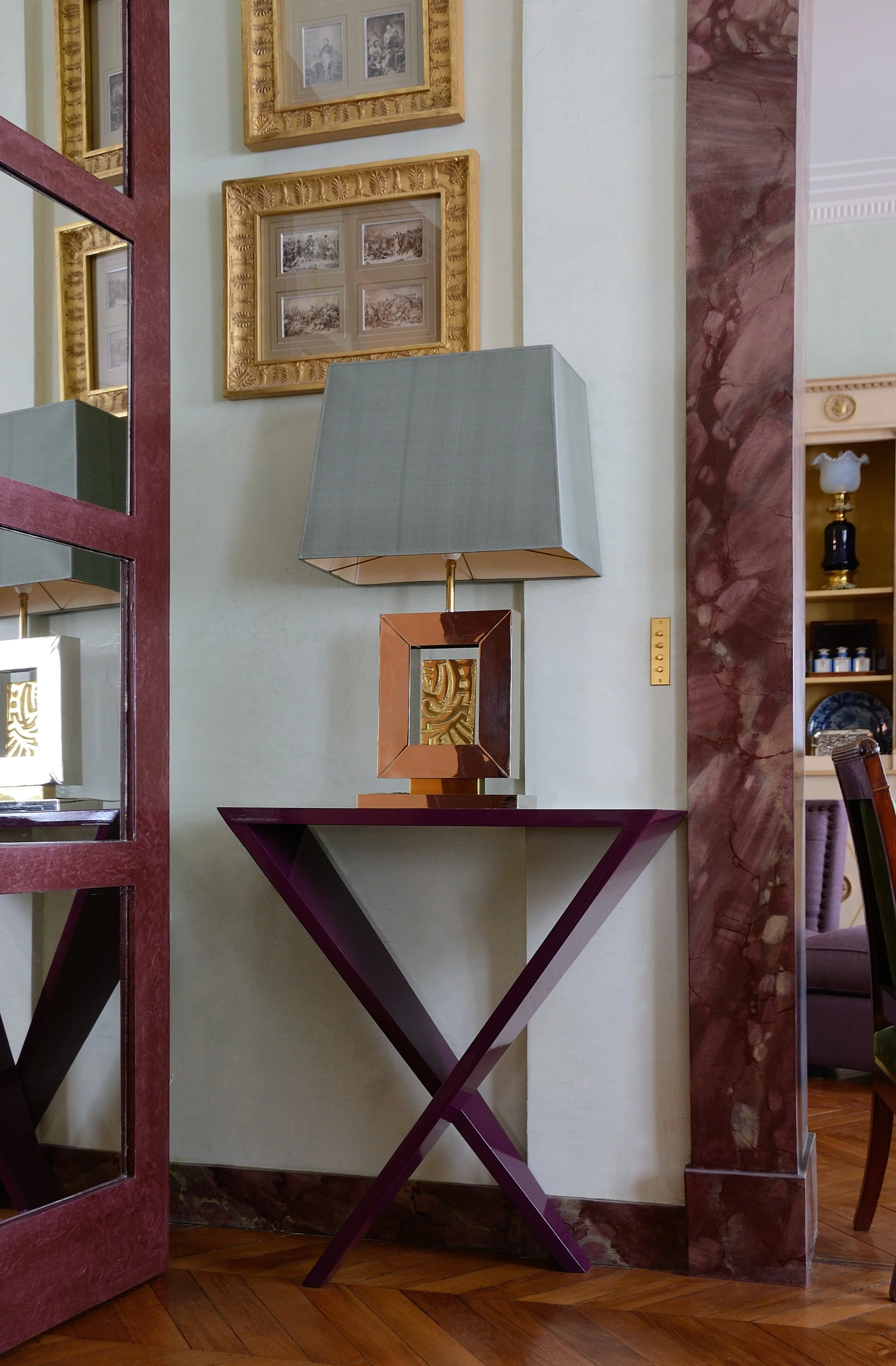 Une lampe de table moderne avec un abat-jour gris clair et une base dorée, placée sur une console violette au pied d'un mur beige avec des cadres dorés contenant des photos en noir et blanc.