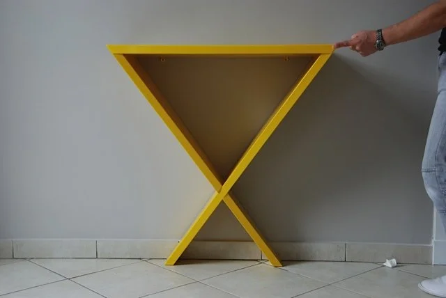 Une table pliée de couleur jaune avec un piétement en X, placée contre un mur blanc, avec une personne tenant le bord droit de la table.