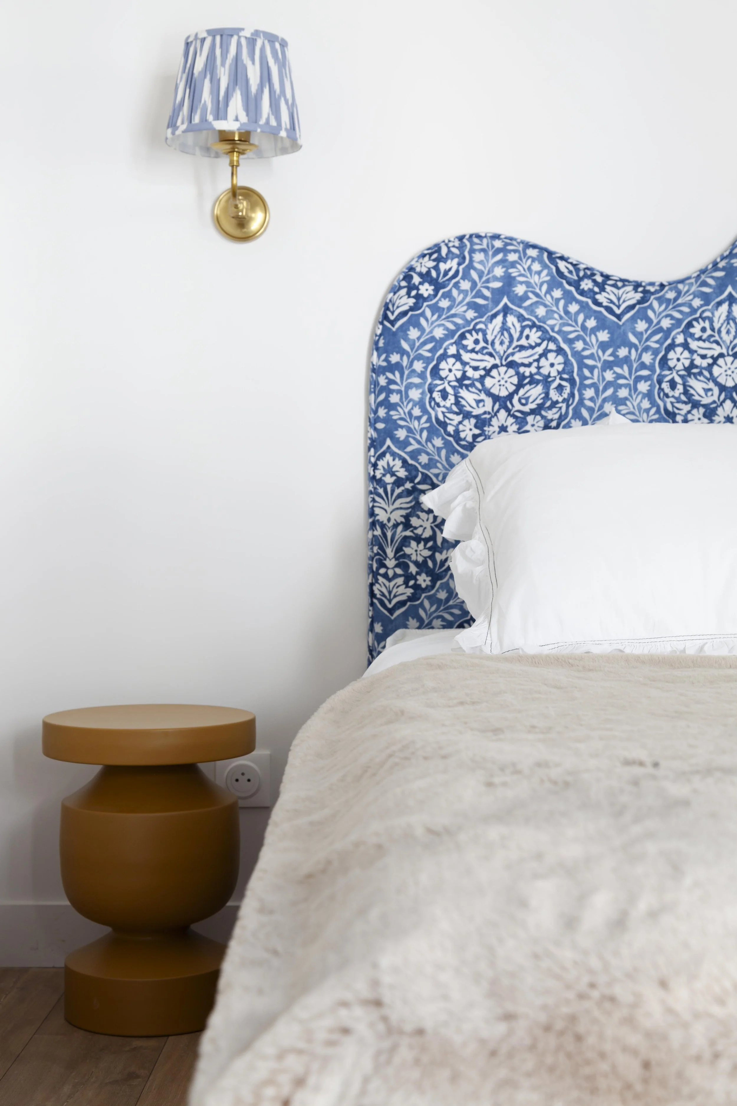Un lit avec une tête de lit bleue ornée d'un motif floral blanc, une table de chevet ronde en bois clair, une lampe à appliqué avec abat-jour bleu et blanc rayé, sur un mur blanc.