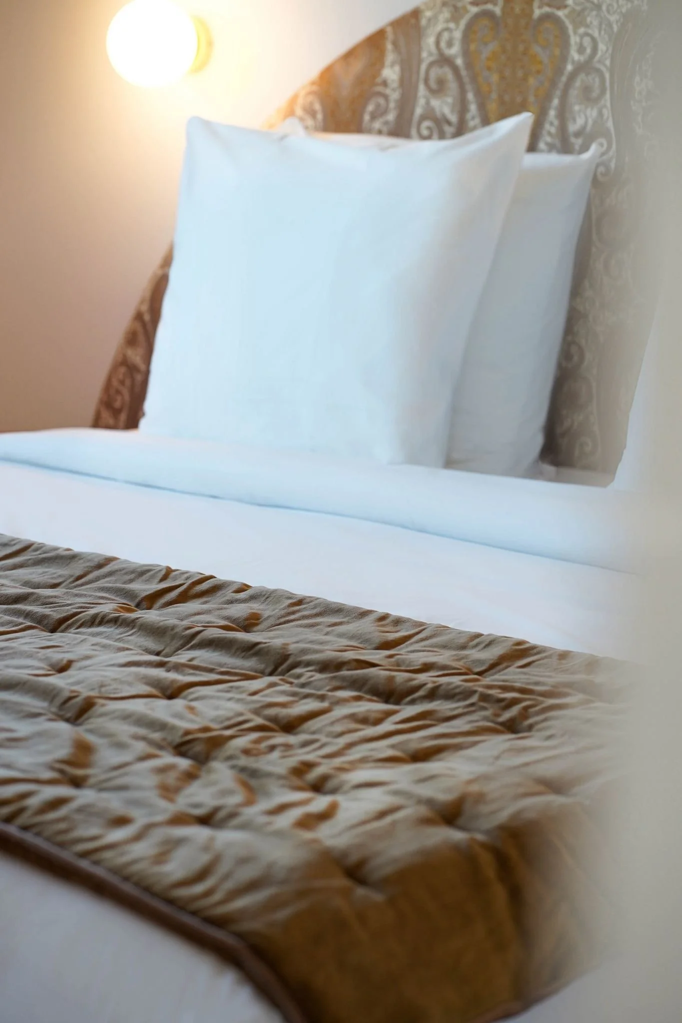 Un lit avec des draps blanc et une couette beige avec motif, sur un chevet en bois, avec deux oreillers blancs, dans une chambre avec un lampadaire au mur et un tête de lit en tissu avec motifs.