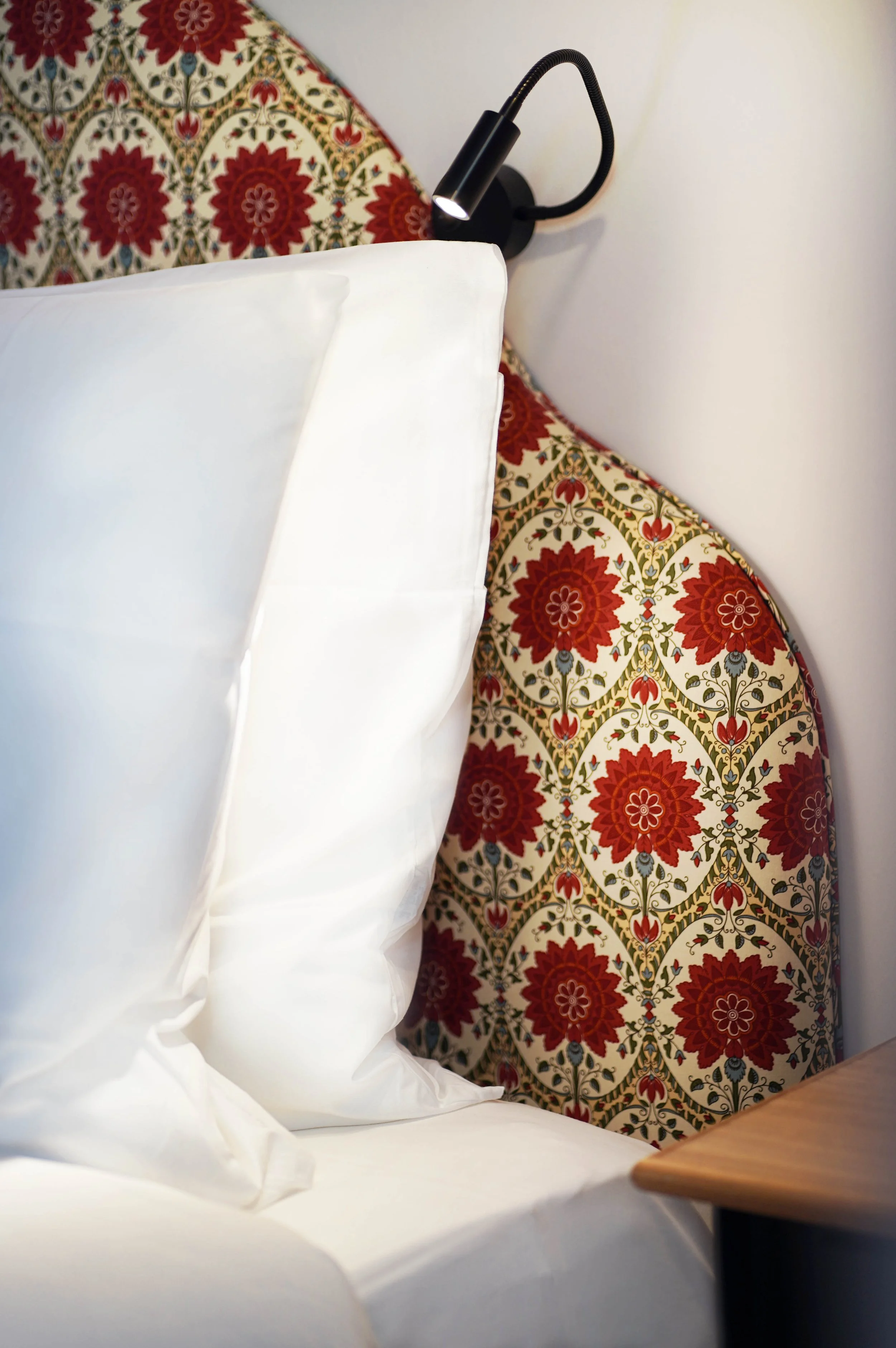 Image d'un lit avec des draps blancs, une tête de lit en tissu avec un motif floral rouge, vert, bleu et beige, sur un mur blanc. Un petit luminaire noir est fixé à la tête de lit.