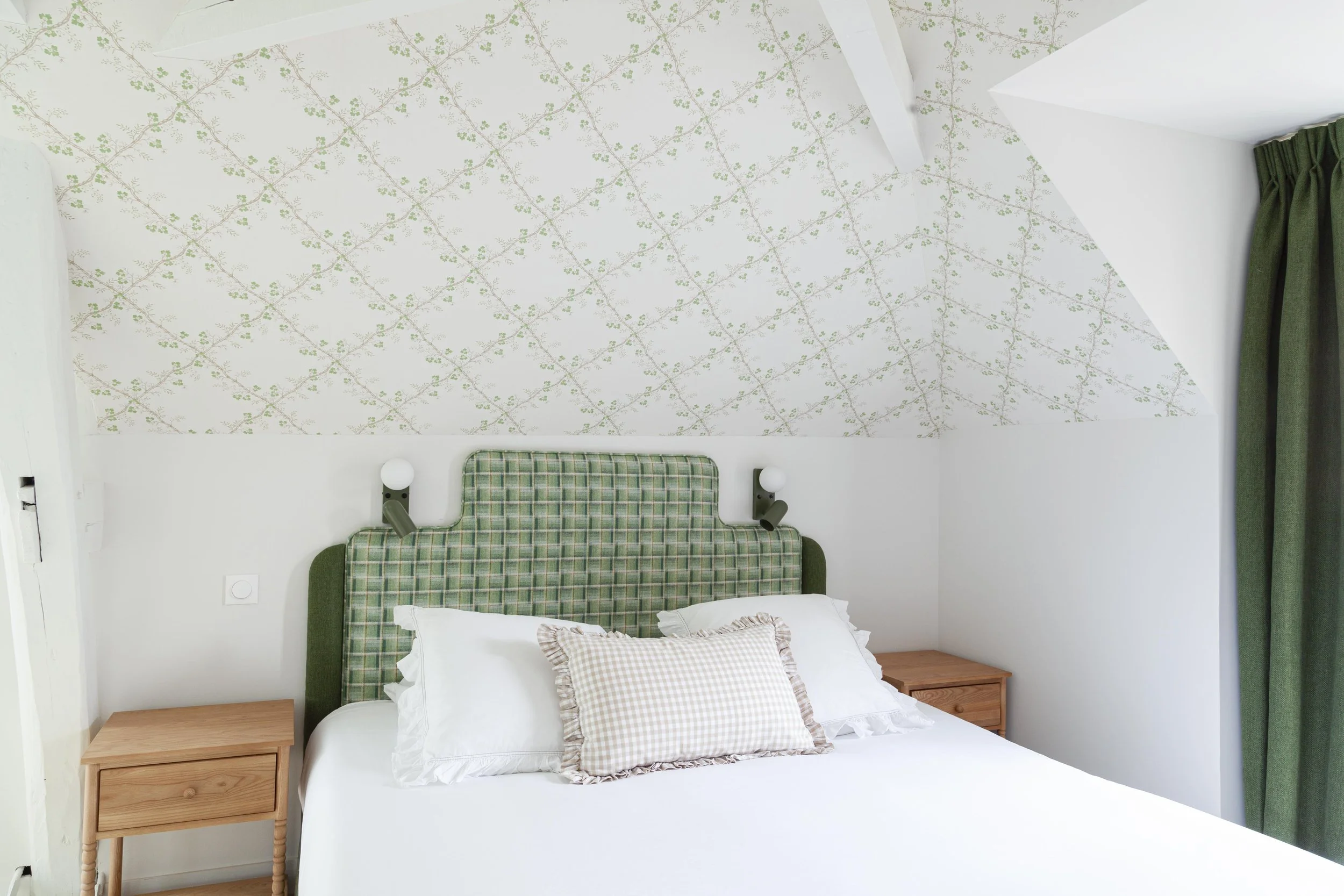 Chambre avec lit double, tête de lit en tissu à carreaux vert et beige, deux tables de chevet en bois naturel, lampe de chaque côté, fenêtre avec rideaux verts, plafond incliné avec papier peint à motif de branches et feuilles vert clair.