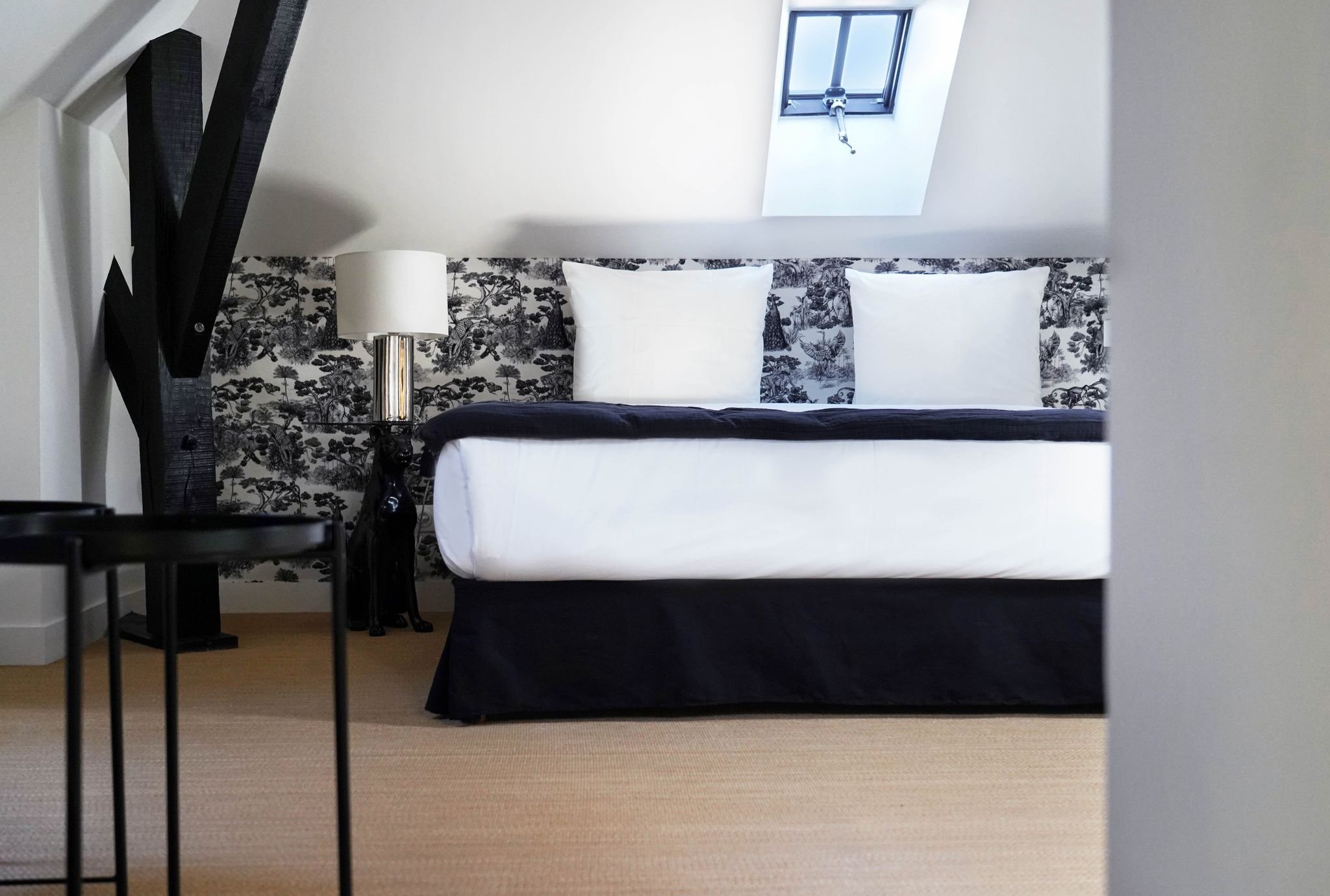 Chambre moderne avec lit à Baldaquin, en arrière-plan un mur tapissé de papier peint noir et blanc avec des motifs de paysages et d'arbres, une lampe et une figurine de chien noir, une fenêtre de toiture.