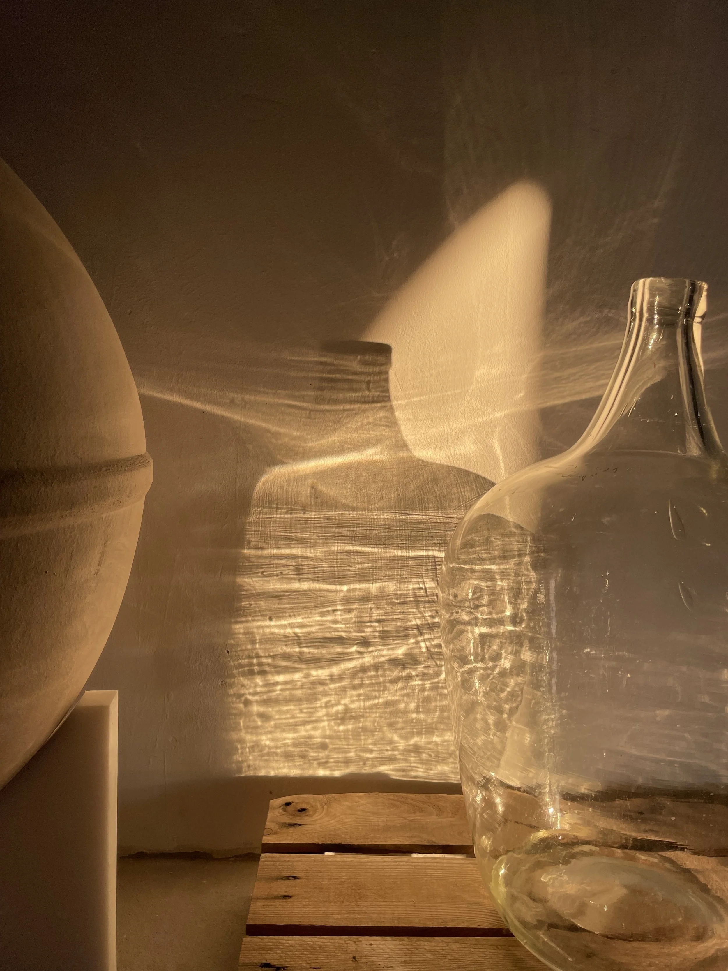 A rustic interior space with warm lighting, featuring a wooden table, a clay pot on the left, a glass carafe on the right, and textured walls with shadows cast by the light.