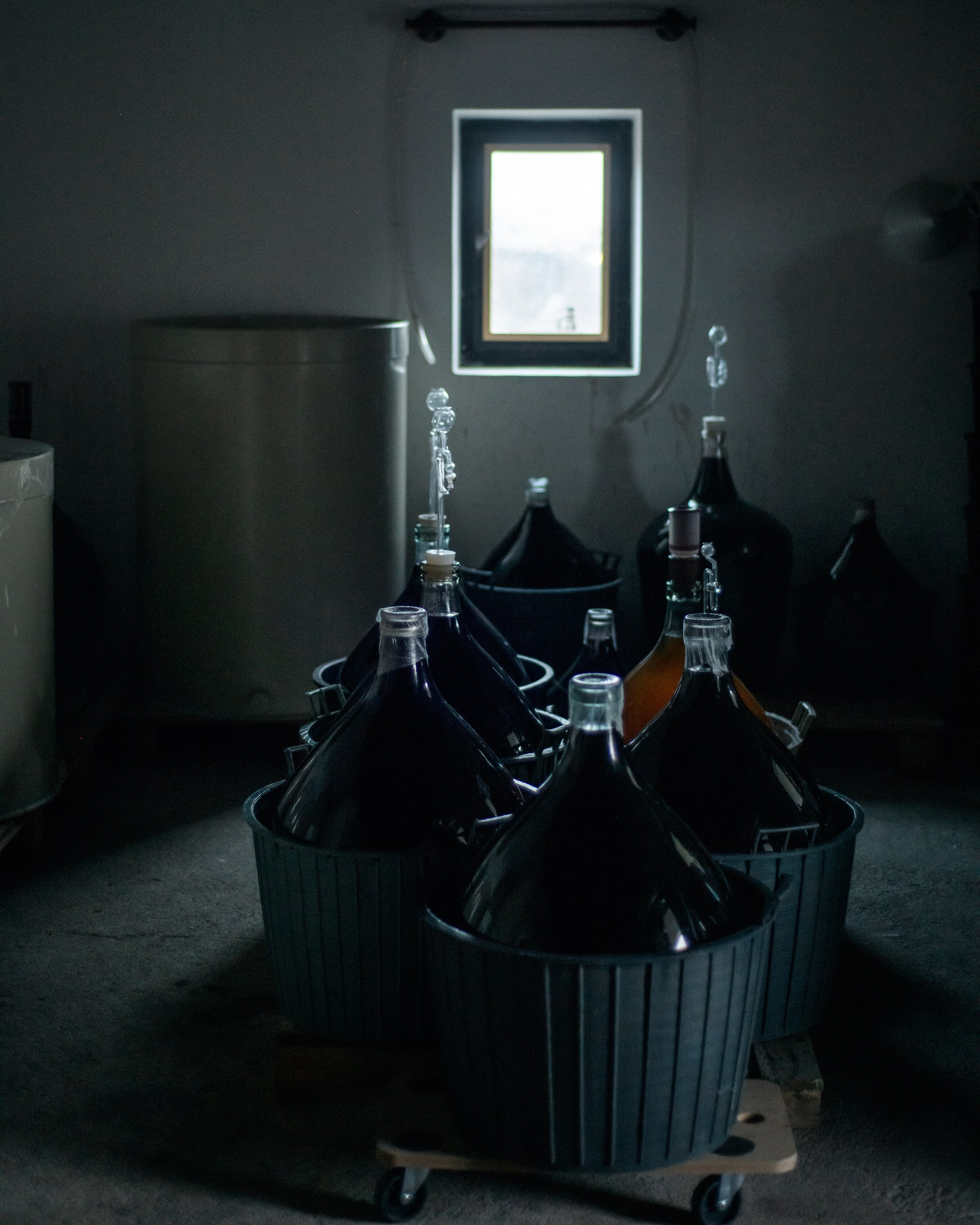 a large gray laundry basket filled with empty glass bottles, placed on a carpeted floor in a dark laundry room with a window in the background.
