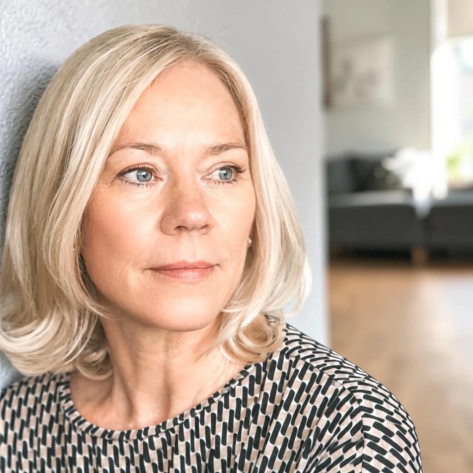 Nahaufnahme einer älteren Frau mit blonden, lockigen Haaren, die nach links schaut, getragen in einem schwarzen und weißen Mustertop, im Hintergrund ein Wohnzimmer mit grauem Sofa und Fenster