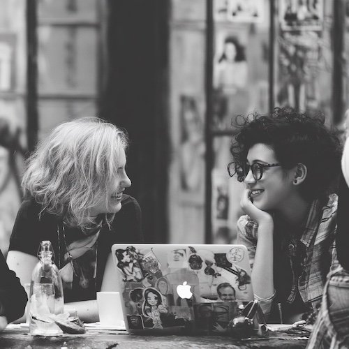 Zwei Frauen lachen und sprechen in einem Café, während ein Laptop mit Stickern davor steht.