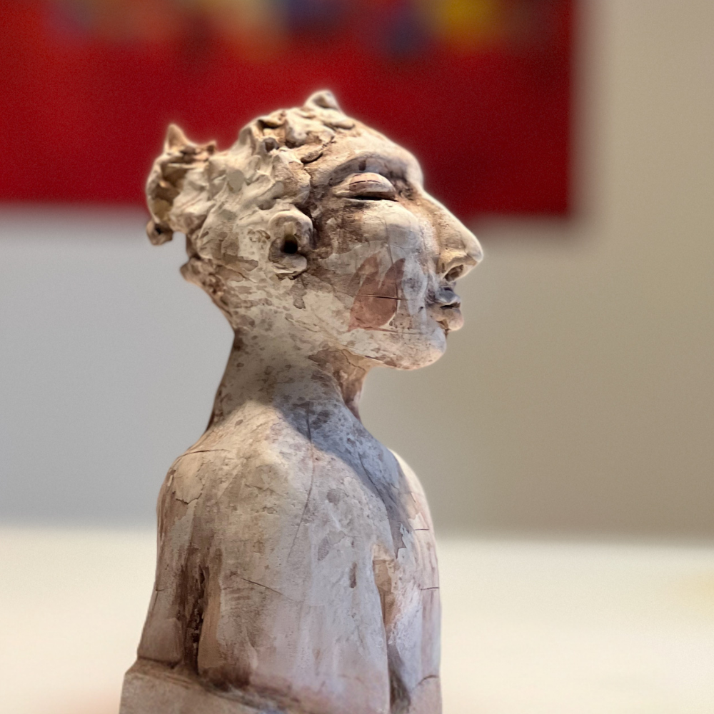 Kunsthandwerkliches Keramikstück, eine Skulptur einer Frau in Profilansicht, mit detailliert ausgearbeiteten Gesichtszügen und Haaren, auf einem weißen Untergrund, mit einem roten Hintergrund im Raum.