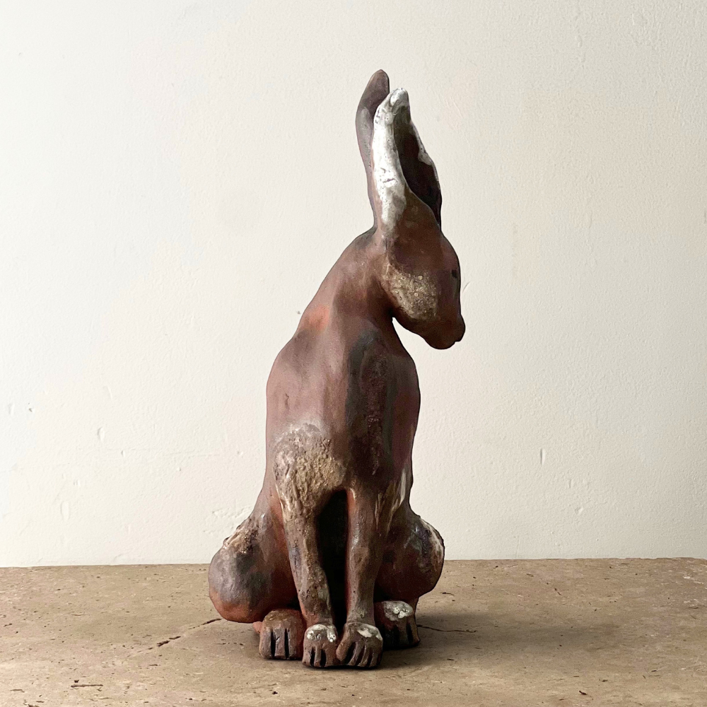 Eine Skulptur eines sitzenden Hundes mit erhobenem Kopf, aus Ton oder Keramik, auf einem Holztisch.