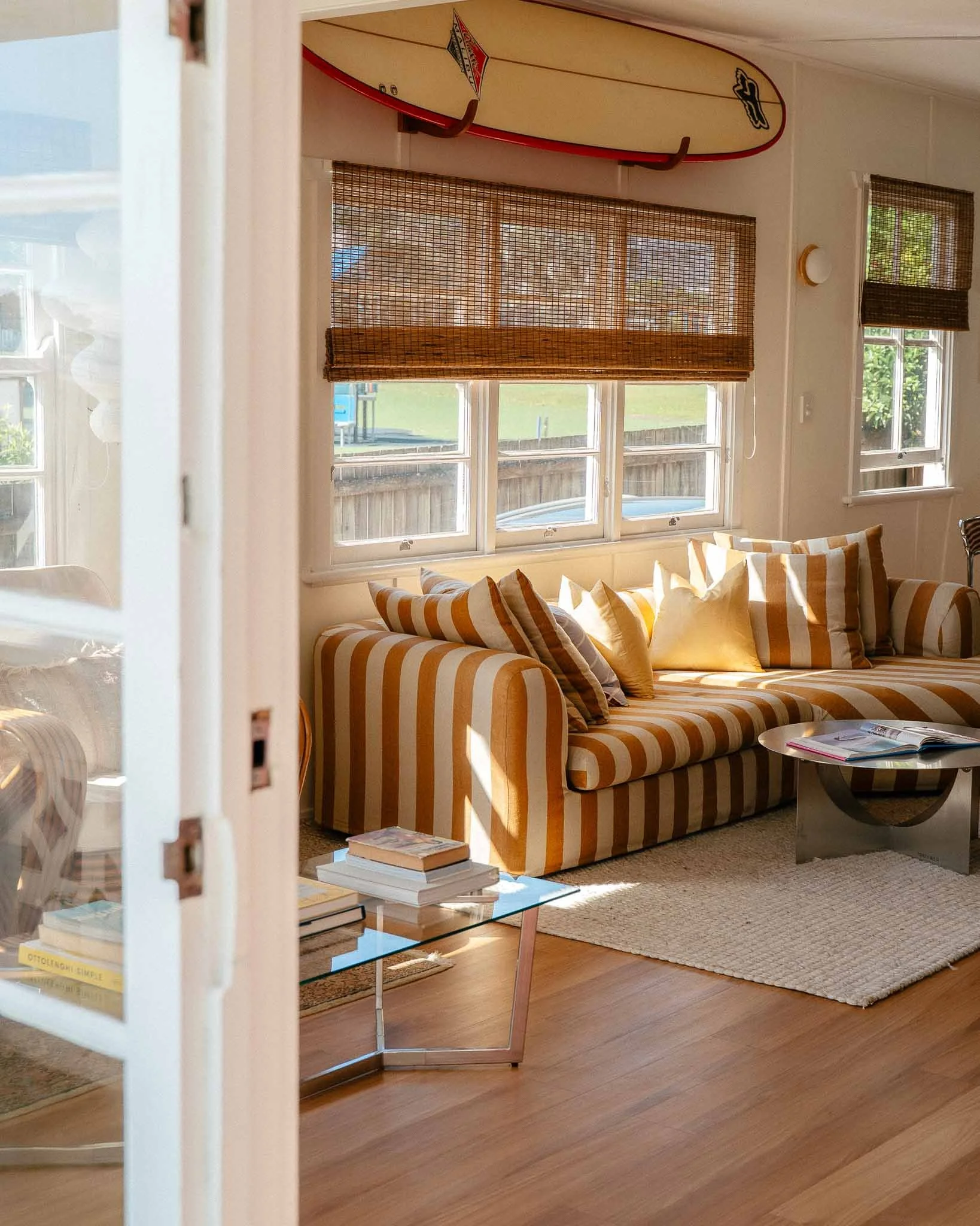 Renovated beach shack living room in Kingscliff, featuring a yellow and white striped sofa, woven blinds, glass coffee table and wall-mounted surfboard.