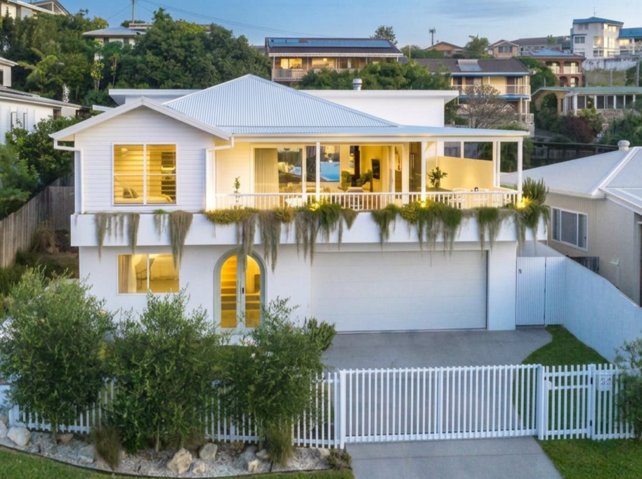 A modern two-story white house with a metal roof, large windows, and a spacious balcony. The house has hanging plants along the balcony and is surrounded by a white picket fence with green bushes in the front yard.
