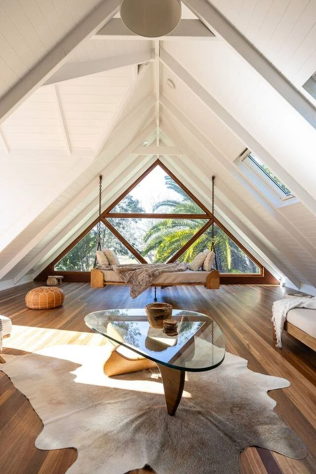 Loft living room in One Palm Building’s Middle Pocket School House renovation, featuring a pitched ceiling, large triangular window overlooking palm trees and a hanging bench swing