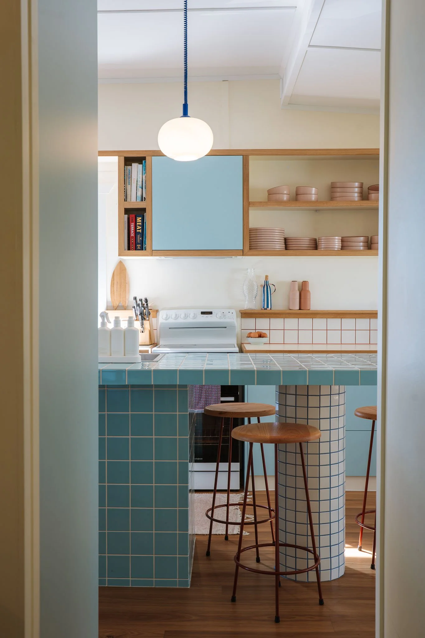 “Kitchen in Holly and Jo’s renovated beach shack, featuring a blue tiled island, timber shelving, white freestanding stove, bar stools and colourful styling.”