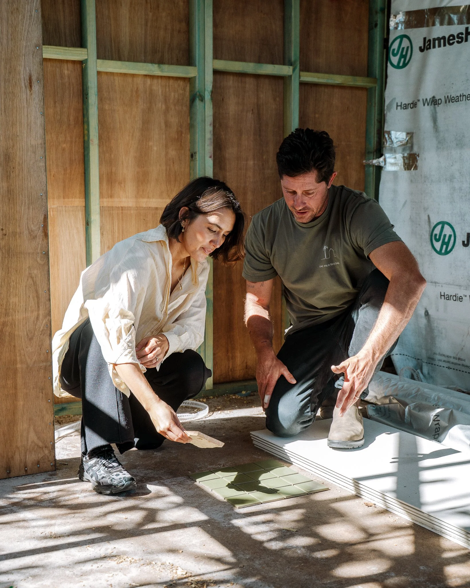 Two people are examining tiles on the ground inside a construction site with wooden framework and insulation around them.