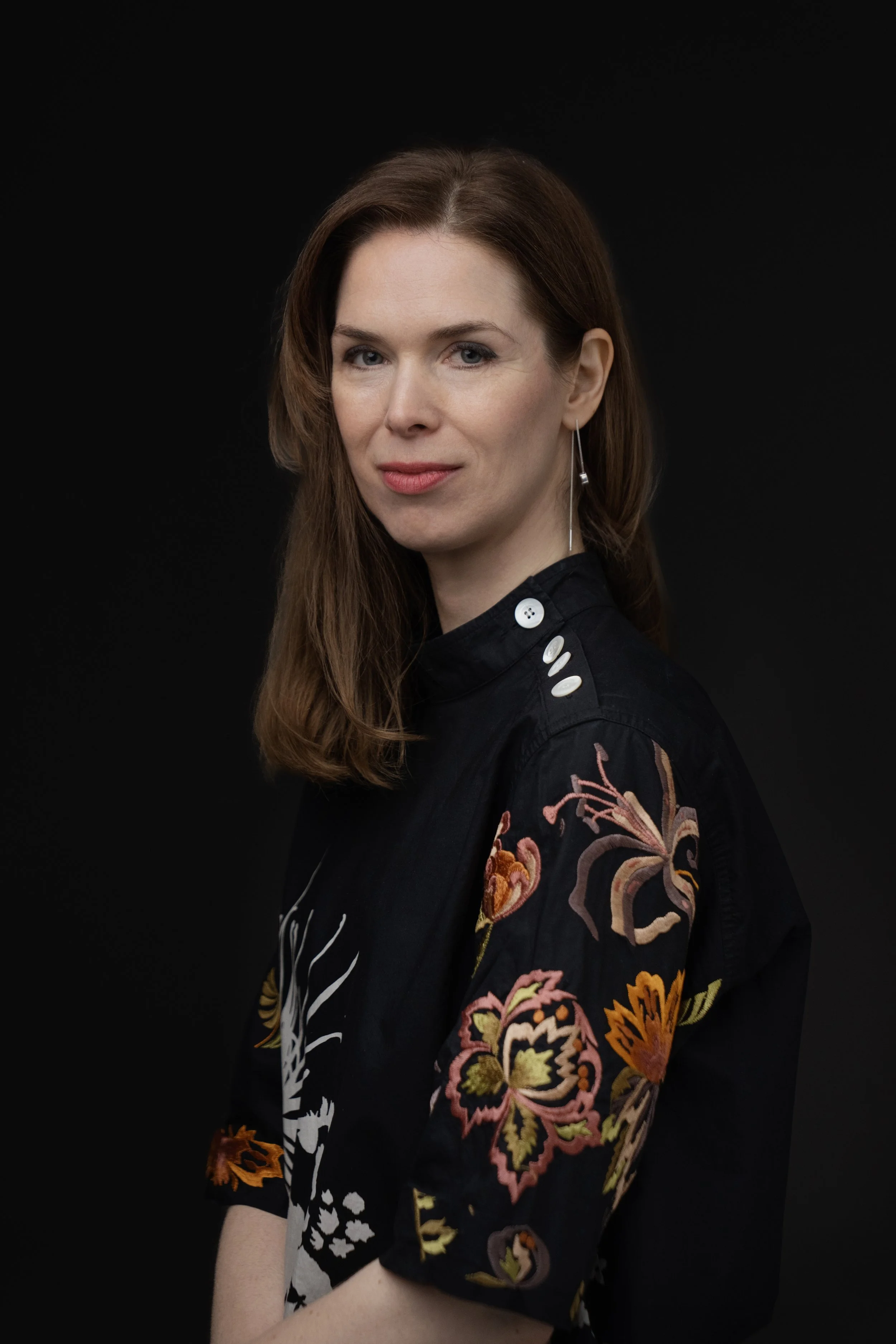 A woman with long brown hair and blue eyes, wearing a black embroidered jacket with floral designs and lapel buttons, standing against a plain black background.
