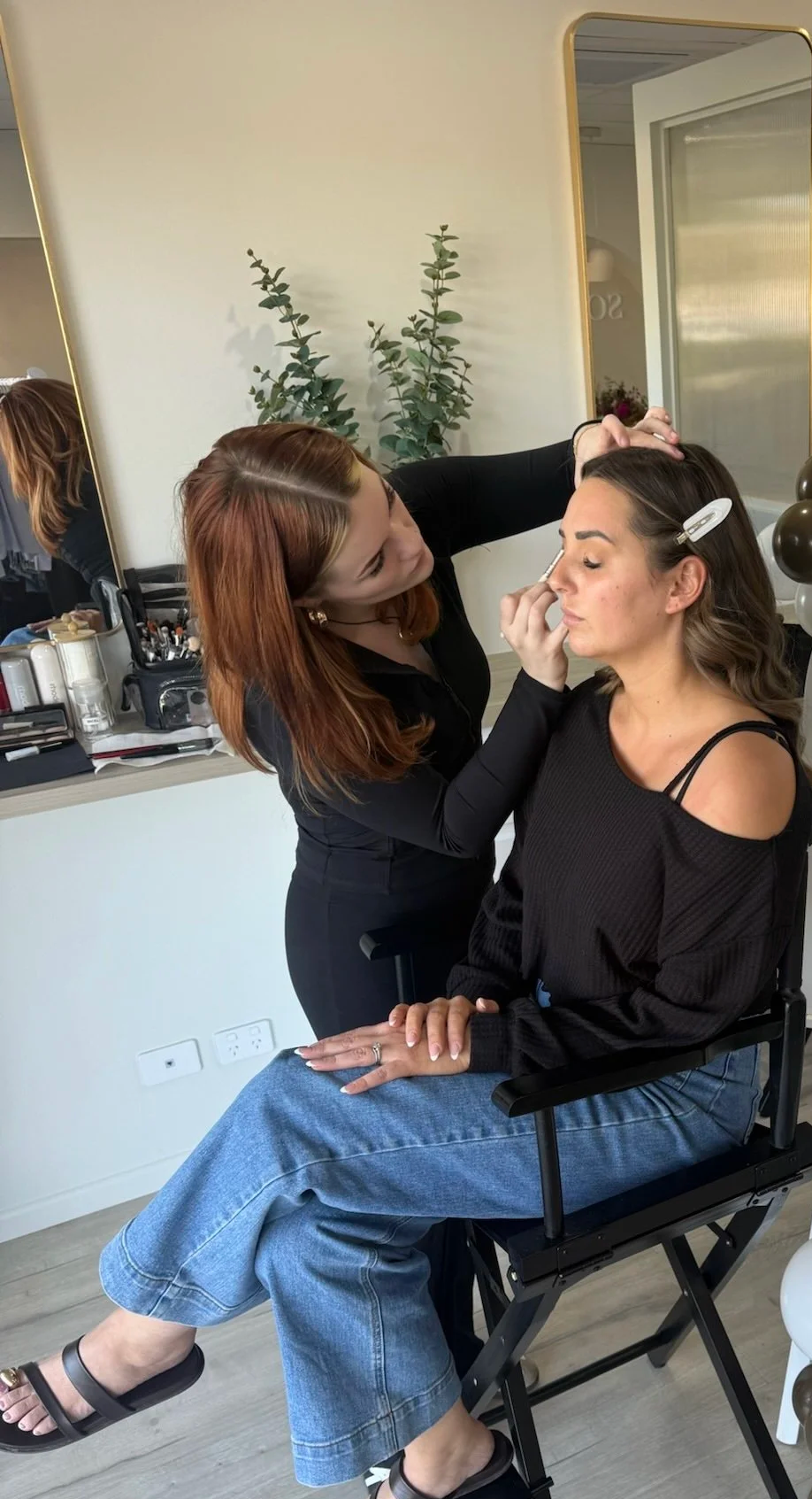 A makeup artist is applying makeup to a woman sitting in a chair, with her eyes closed. The woman is wearing a black top and blue jeans. The setting appears to be a makeup studio with a mirror, makeup supplies, and some greenery in the background.