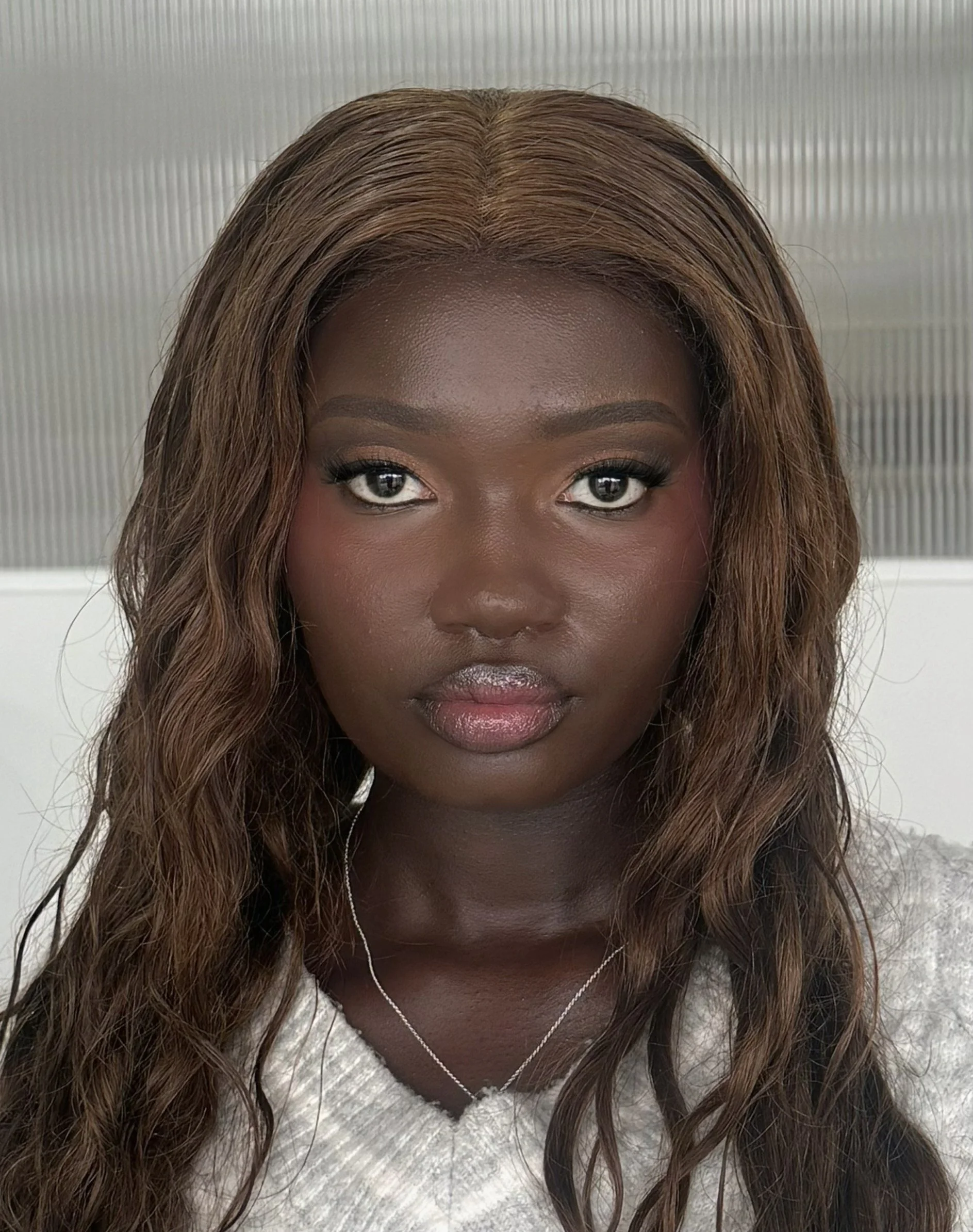 Close-up of a woman's face with polished lips, makeup, and long wavy hair. She's wearing a white fuzzy sweater and a silver necklace, standing in front of a textured metallic background.